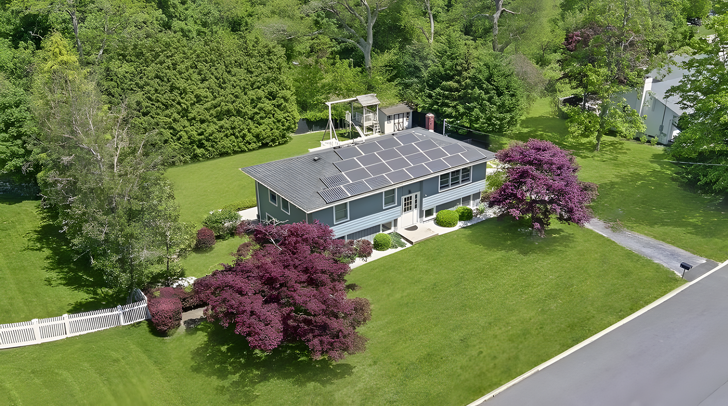A house with a dark gray roof covered in solar panels situated in a lush green yard with purple flowering trees and a white picket fence.