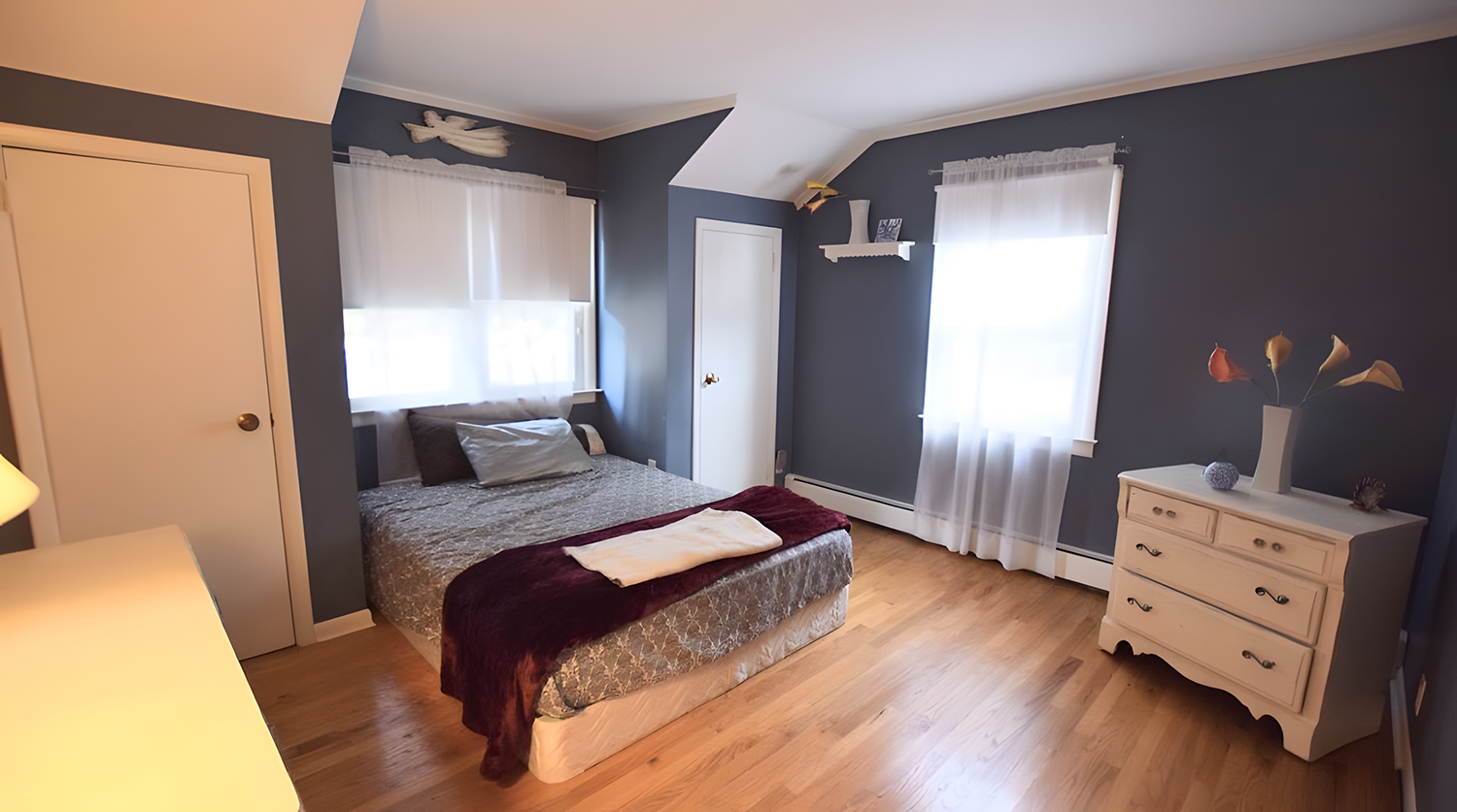 Bedroom with blue walls, white curtains, a bed with gray bedding and pillows, a white dresser with a vase and decorative items, and wooden flooring.