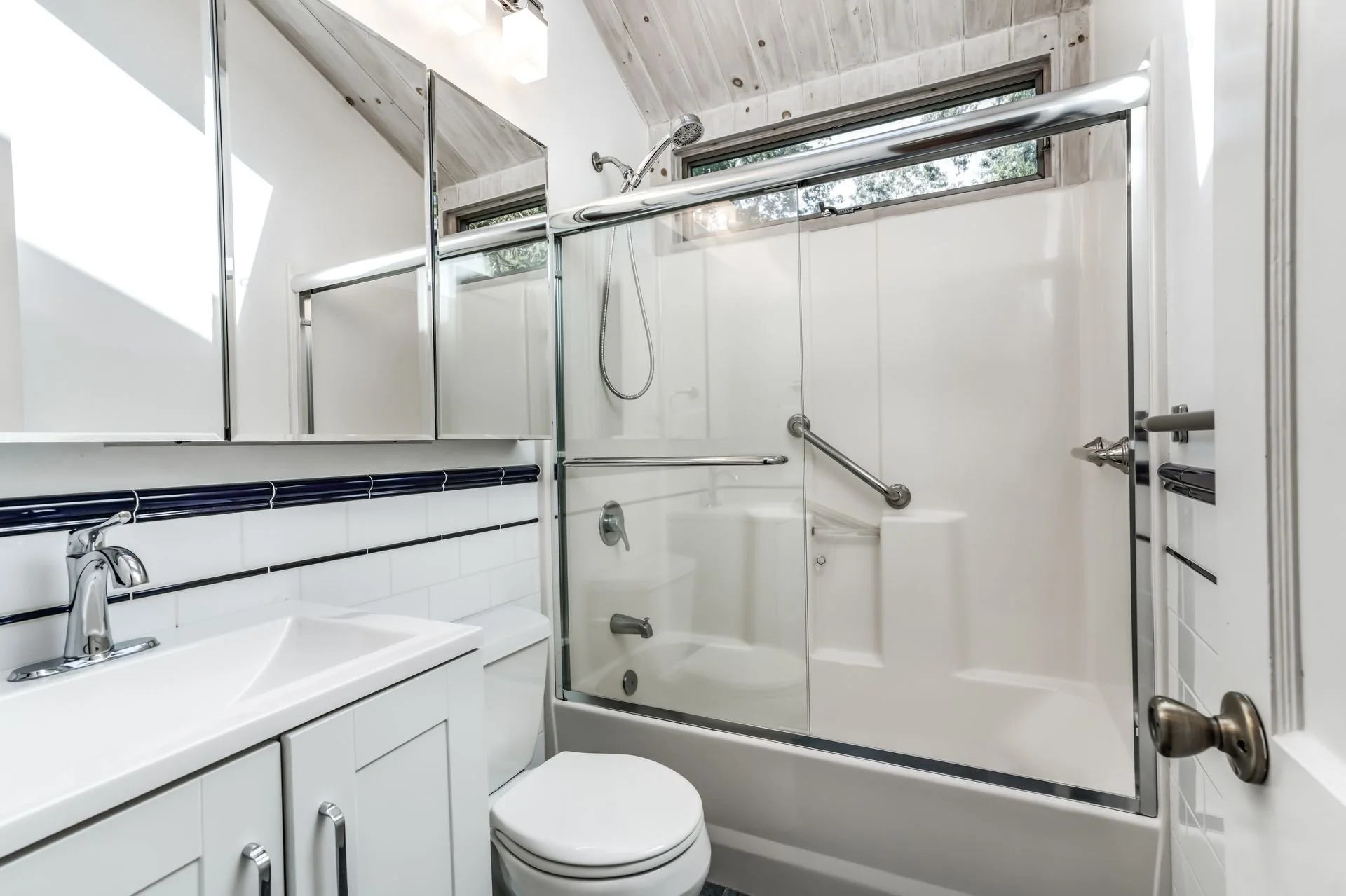 Small bathroom with white tiled walls, a toilet, a sink with a cabinet, and a shower with a glass door and metal handrails. There are two narrow windows near the ceiling and ceiling-mounted light fixtures.