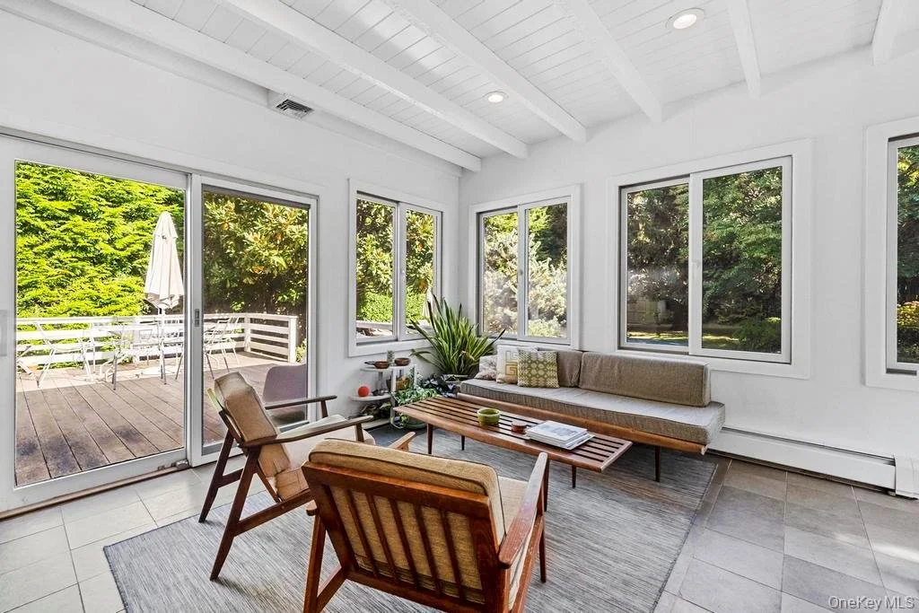 Bright sunroom with beige seating, wooden coffee table, and large windows showing a lush green backyard with trees and a wooden deck.