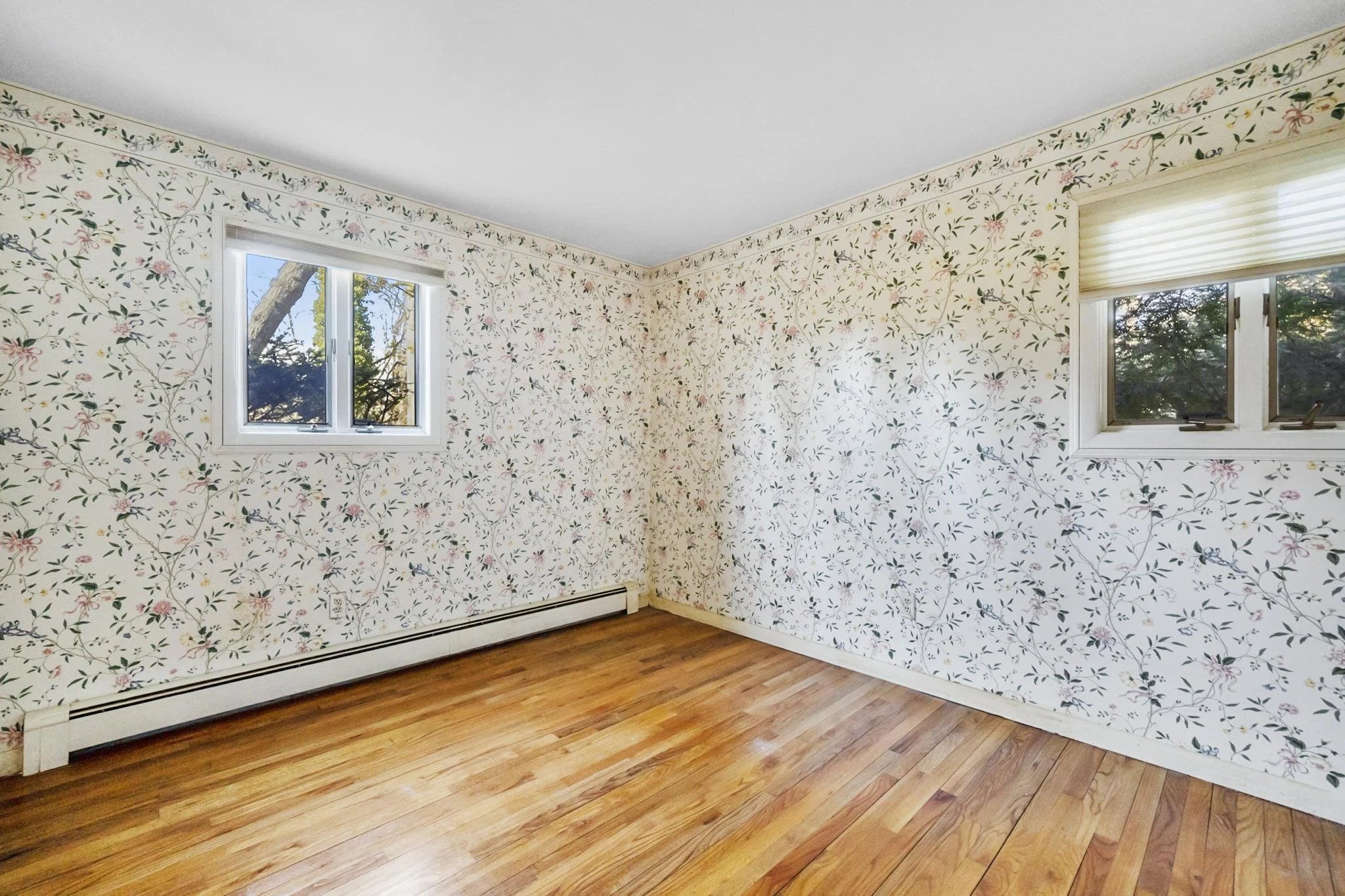 Empty room with floral wallpaper, hardwood floors, two windows, and baseboard heating