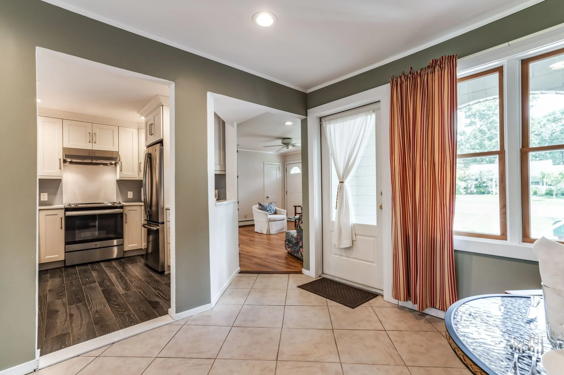 Kitchen and living room area with large windows and a door to the outside. The kitchen has white cabinets and stainless steel appliances. The living room has a white chair, a ceiling fan, and a door with a white curtain. There are striped red and yel