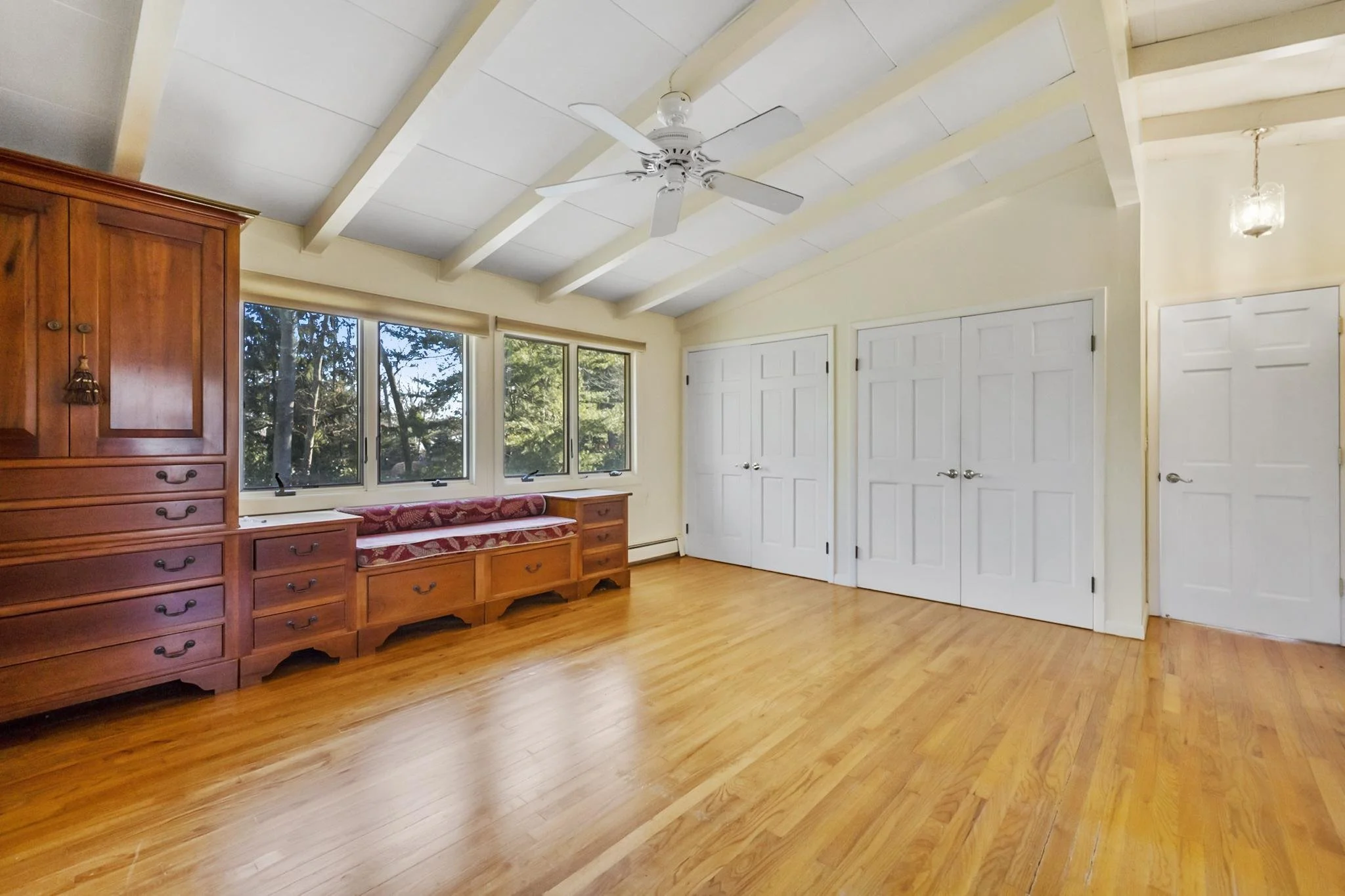 Empty room with wooden floors, white walls, large windows, a ceiling fan, double doors, a built-in wooden cabinet, and a small bench with a cushioned seat.