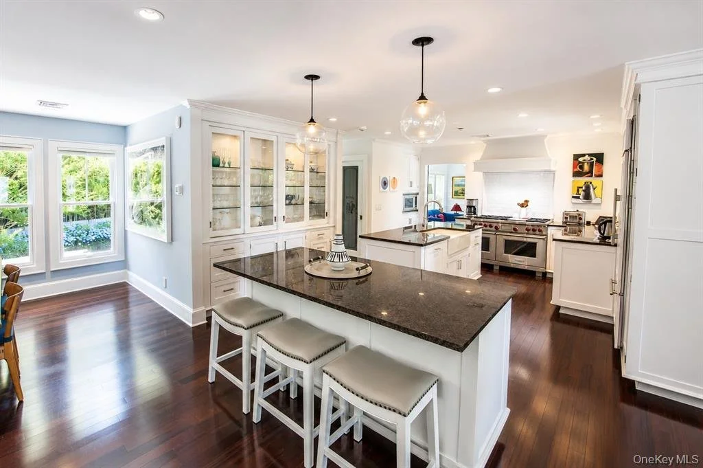 Bright, spacious kitchen with white cabinetry, dark granite countertops, hardwood floors, and a large island with seating and pendant lighting. There are windows with views of greenery outside, and a display cabinet with glass doors.