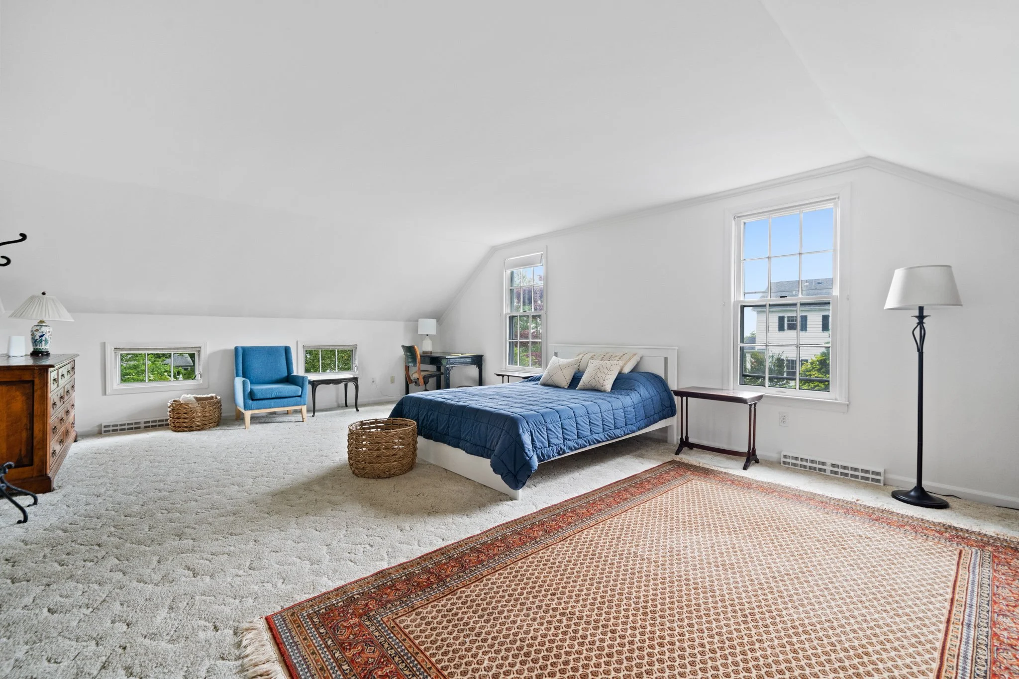 A spacious bedroom with white walls and large windows letting in natural light, featuring a bed with dark blue quilt and several pillows. The room includes a beige carpet with a patterned rug, a blue armchair, a small black desk with a lamp, and wick