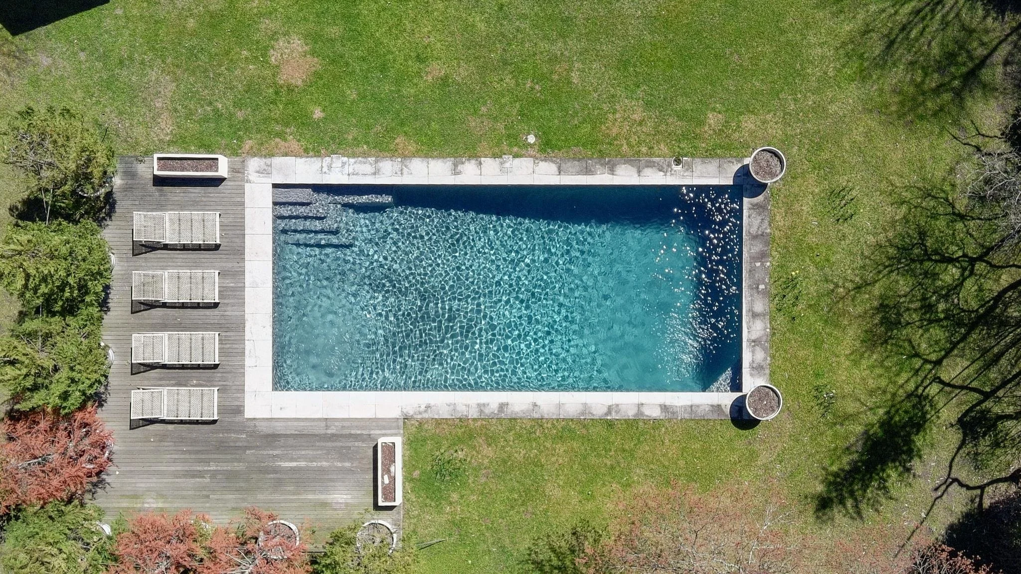 Aerial view of a rectangular swimming pool with blue water, surrounded by a paved deck with lounge chairs, adjacent to a grassy area with trees and shrubs.