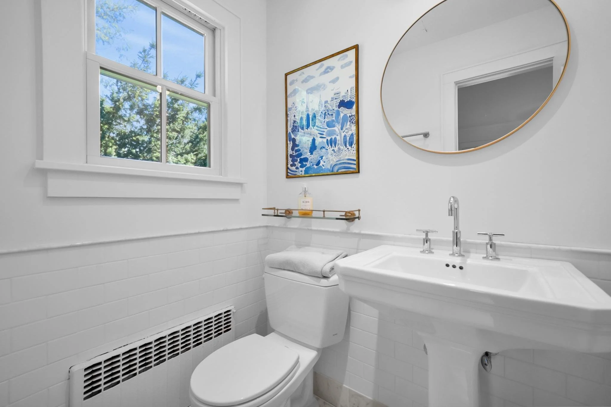 A small white bathroom with a window showing green trees outside, a framed blue and white abstract artwork, a round mirror with a gold rim, a white sink, and a toilet with a white towel on top. There is a vent below the window.
