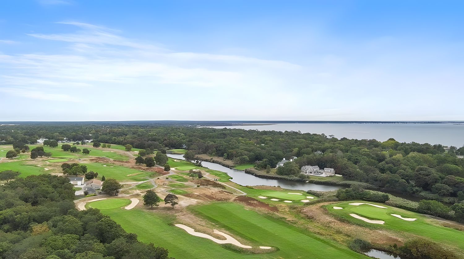 Aerial view of a golf course near a body of water, with green fairways, sand bunkers, trees, and houses, under a partly cloudy sky.