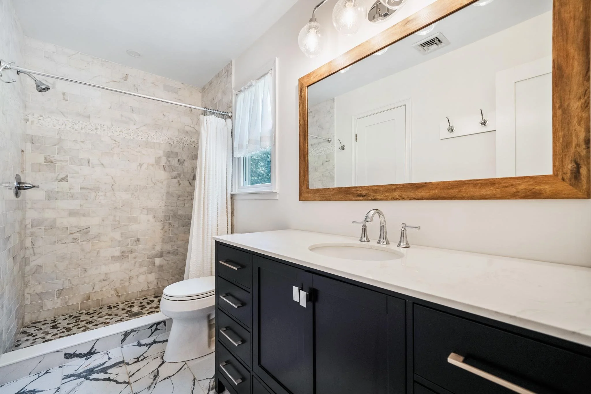 Modern bathroom with a walk-in shower, marble tile floor, vanity with black cabinets, white countertop, large mirror, and a window with sheer white curtains.