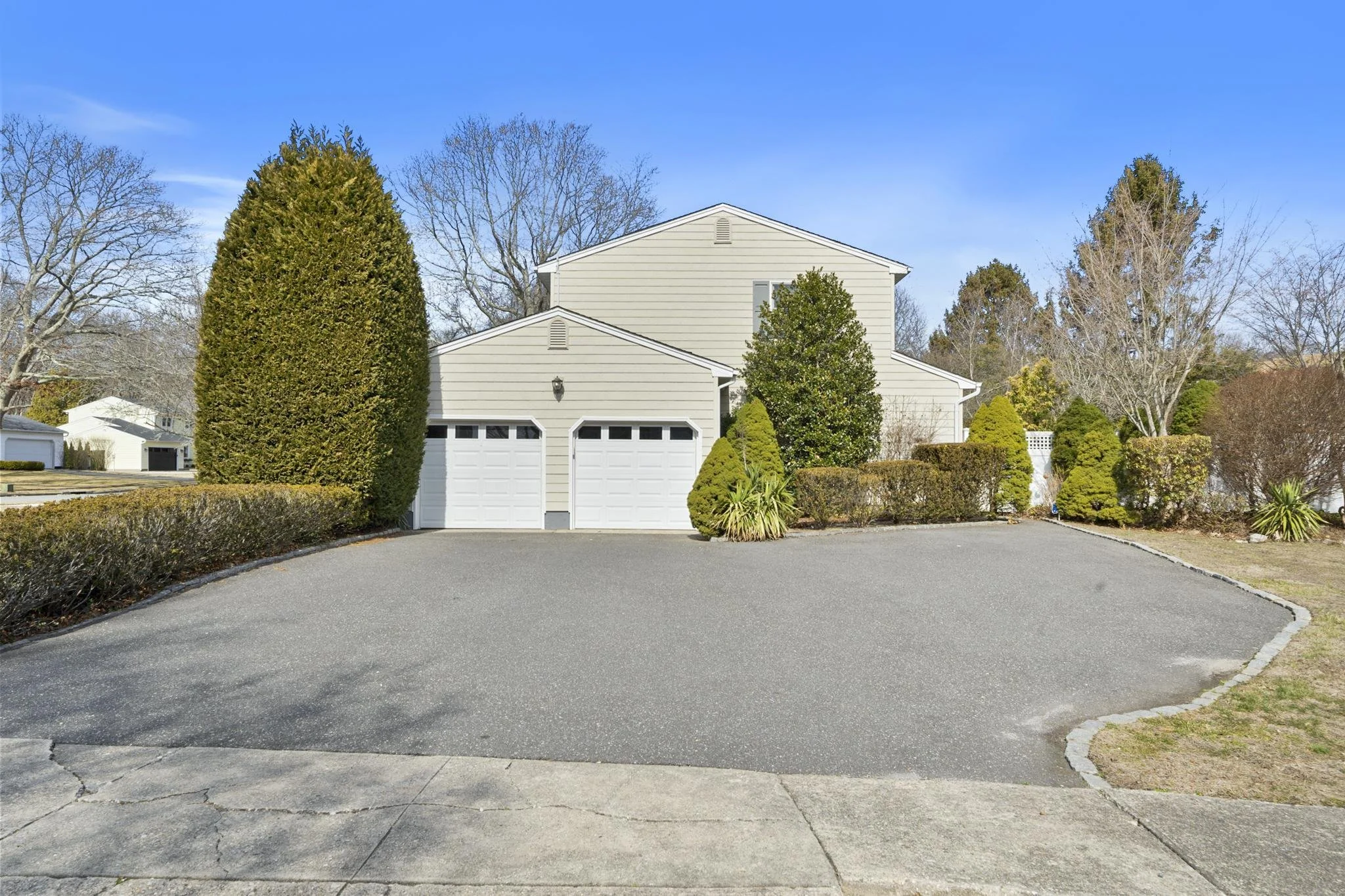 Front view of a beige house with double garage, surrounded by trees and bushes, under a clear blue sky.