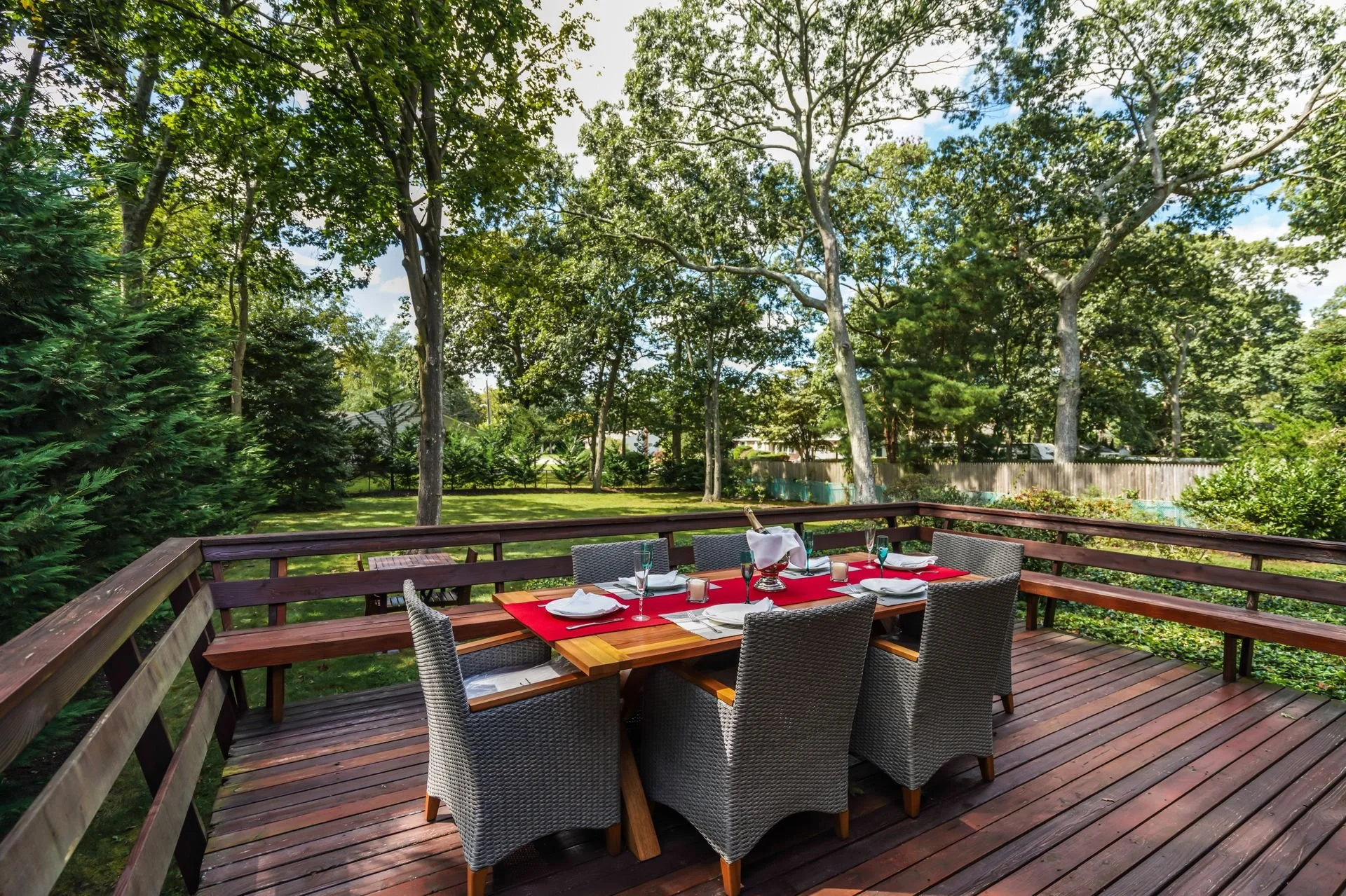 A backyard deck with a dining table set for a meal, surrounded by trees and a wooden fence.