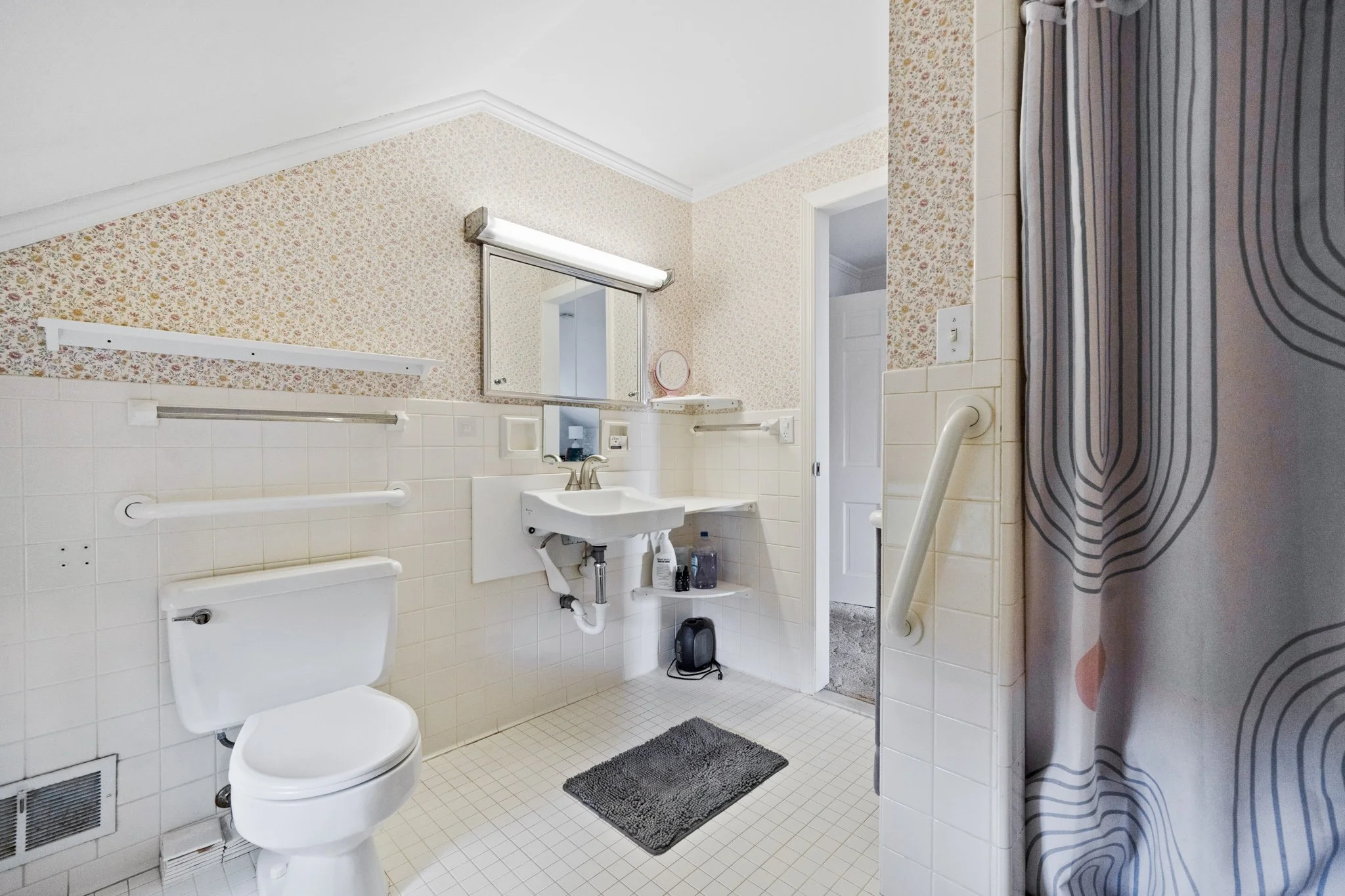 A small bathroom with a toilet, a sink, a mirror, and a shower curtain with a swirling pattern. The walls have floral wallpaper and white tiles. There is a gray rug on the floor and some toiletries near the sink.