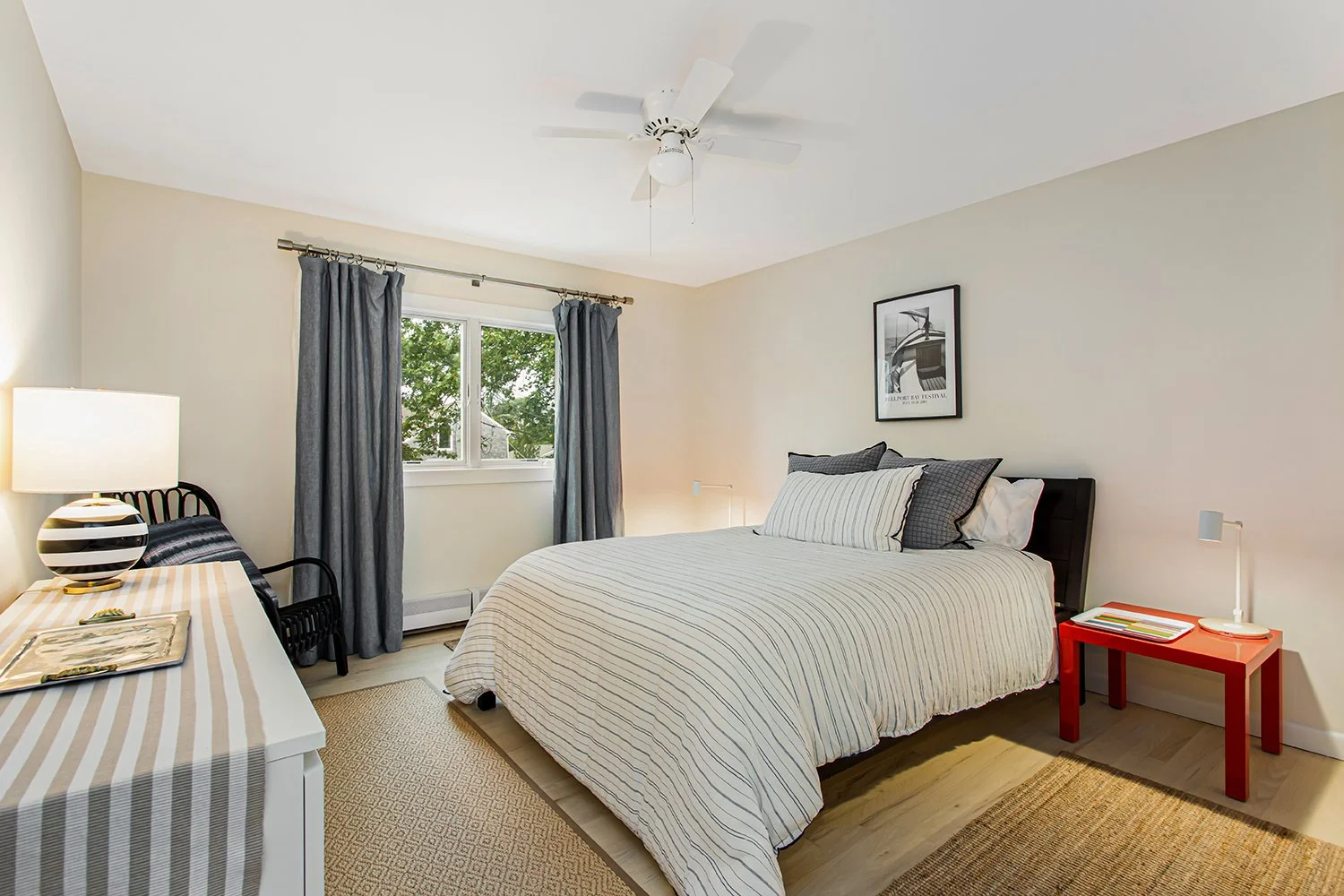 A bedroom with a bed, striped bedding, a red side table, a black chair, a table with a striped lamp, and a window with blue curtains, beige walls, and hardwood floors.