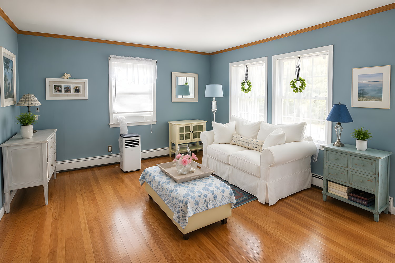 Living room with blue walls, hardwood floor, white sofa, two side tables with lamps, blue and white ottoman, windows with curtains and hanging wreaths, potted plants, framed pictures, and a small air conditioner.