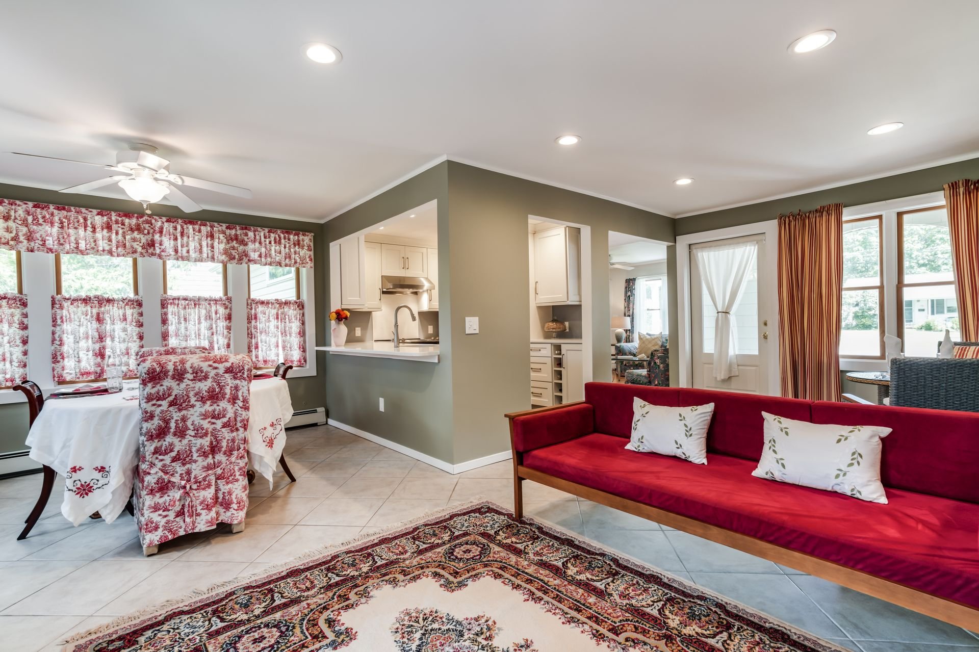 Bright living room with green walls, white ceiling, and recessed lighting. Features a red sofa with pillows, a dining table with chairs covered in red and white toile fabric, a large patterned area rug, and windows with red and white curtains. A kitc