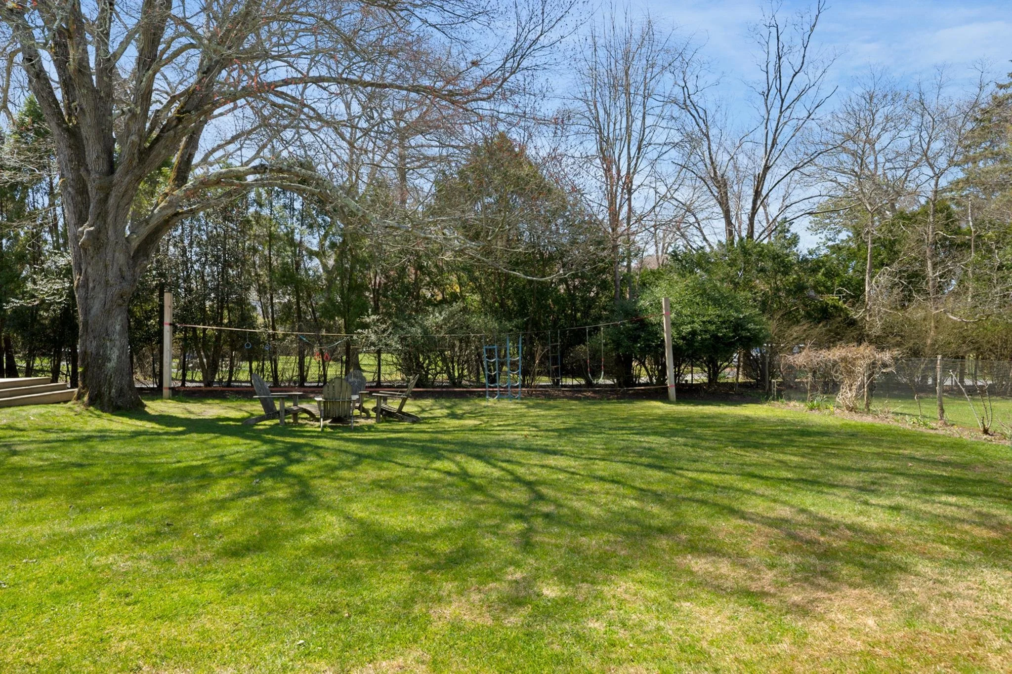 A backyard scene with a large tree, a grassy lawn, outdoor chairs, and a playset with swings. Several leafless trees are in the background, and the sky is clear and blue.