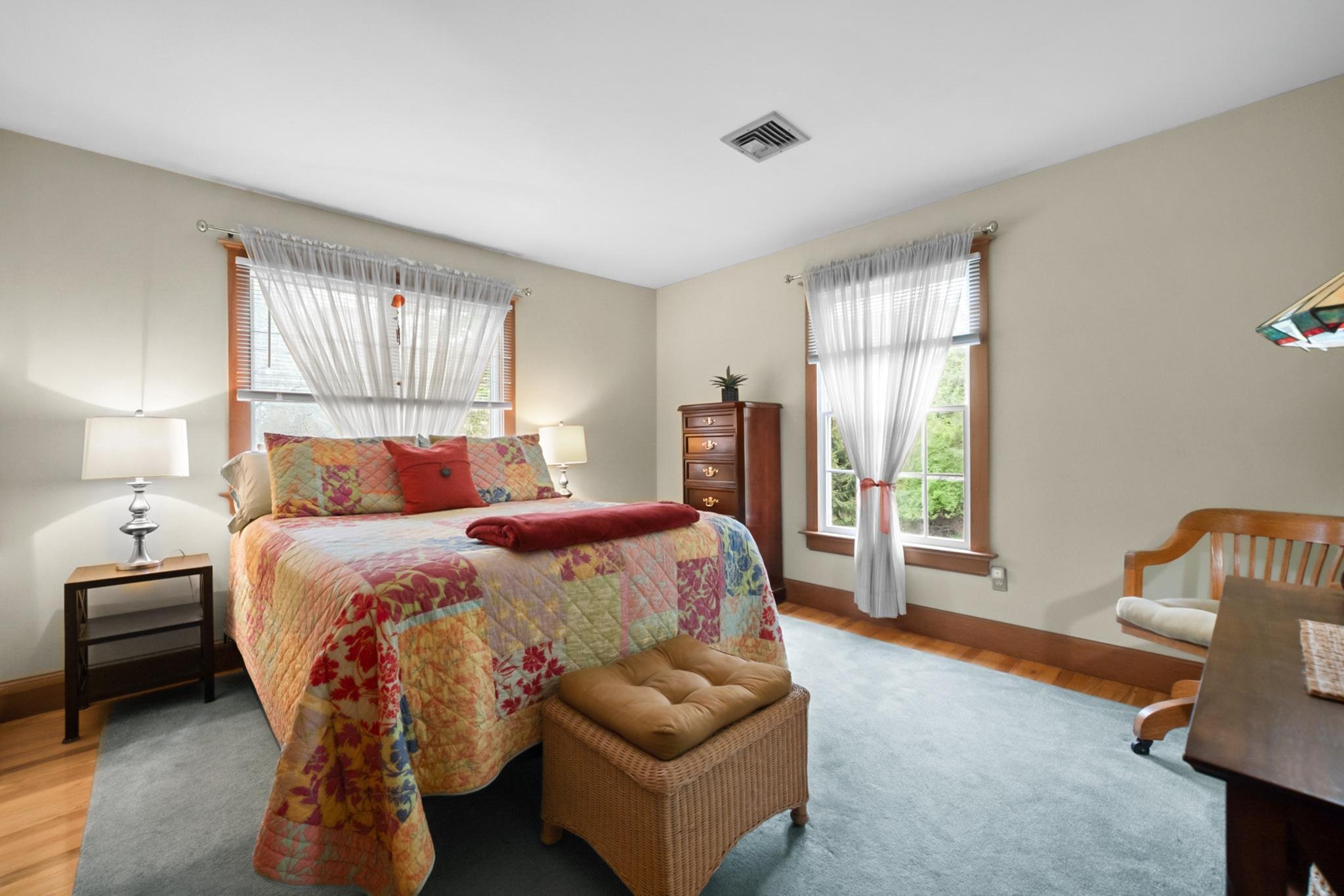 A cozy bedroom with a colorful quilted bedspread, pillows, and a blanket, with two nightstands and lamps on each side of the bed, and two windows with sheer white curtains letting in natural light.