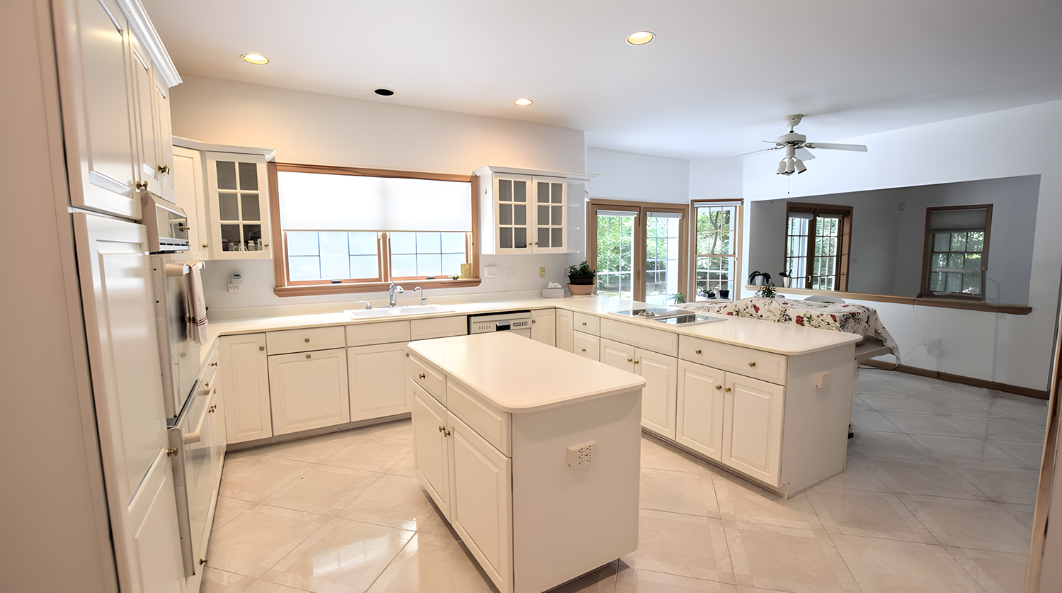 Bright white kitchen with island, cabinets, large windows, and a view into a dining area with a table and chairs.