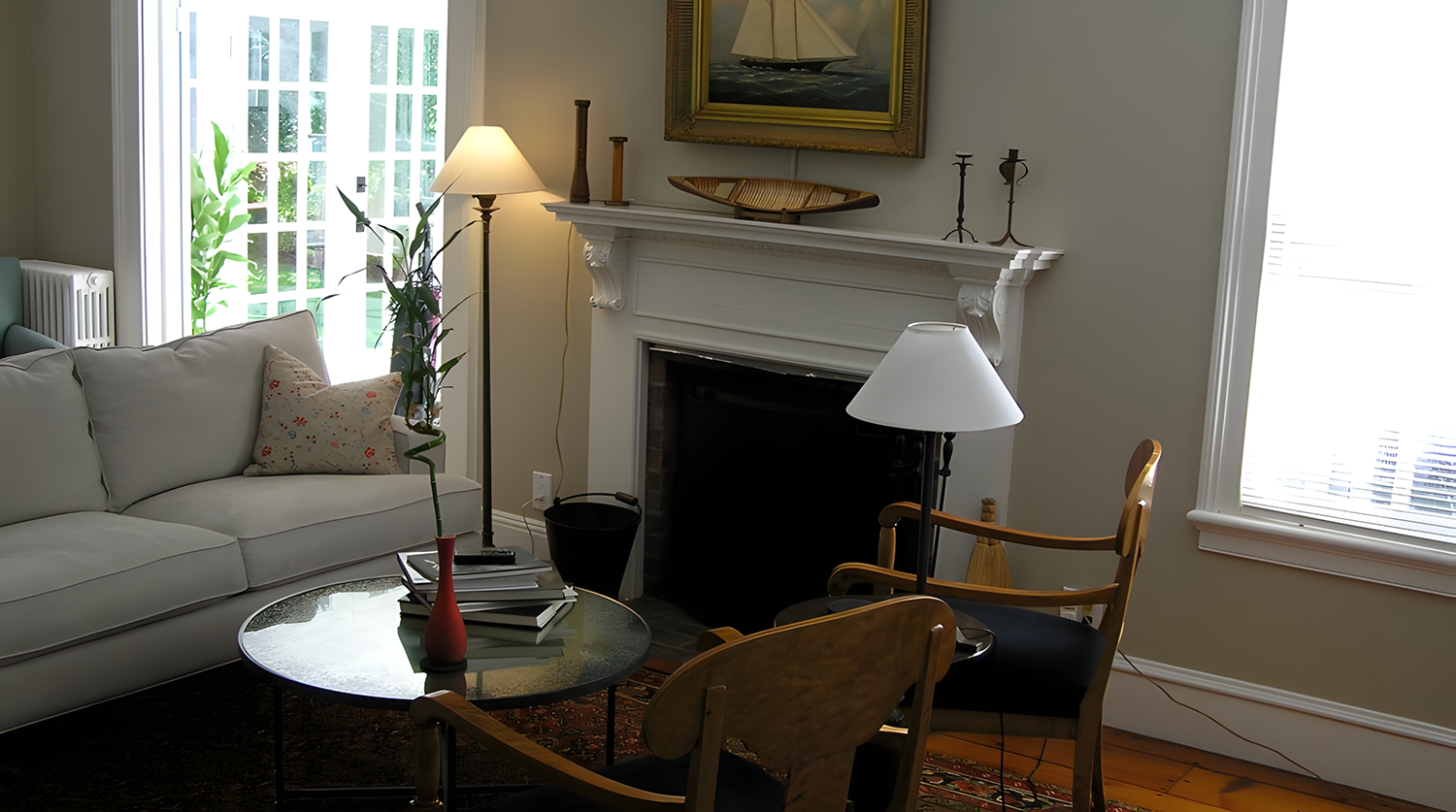 Living room with a white couch, a glass coffee table with a red vase and books, a fireplace with decorative items, a wooden armchair with a white lampshade, and window blinds letting in natural light.