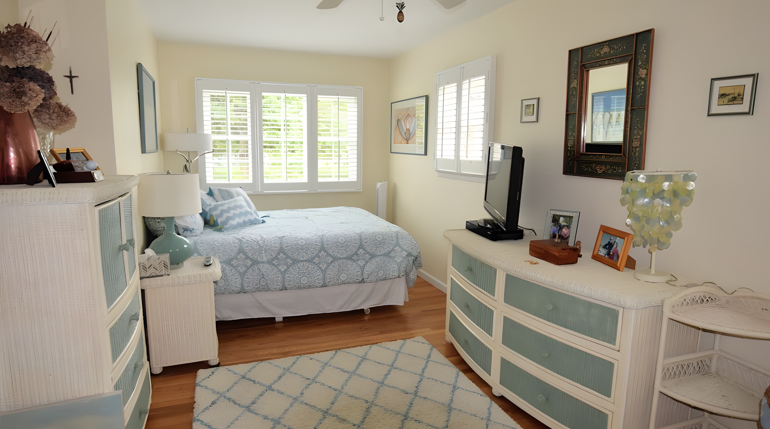A cozy bedroom with a bed, wicker furniture, framed artwork, a TV, and various decorative items near windows with white plantation shutters.