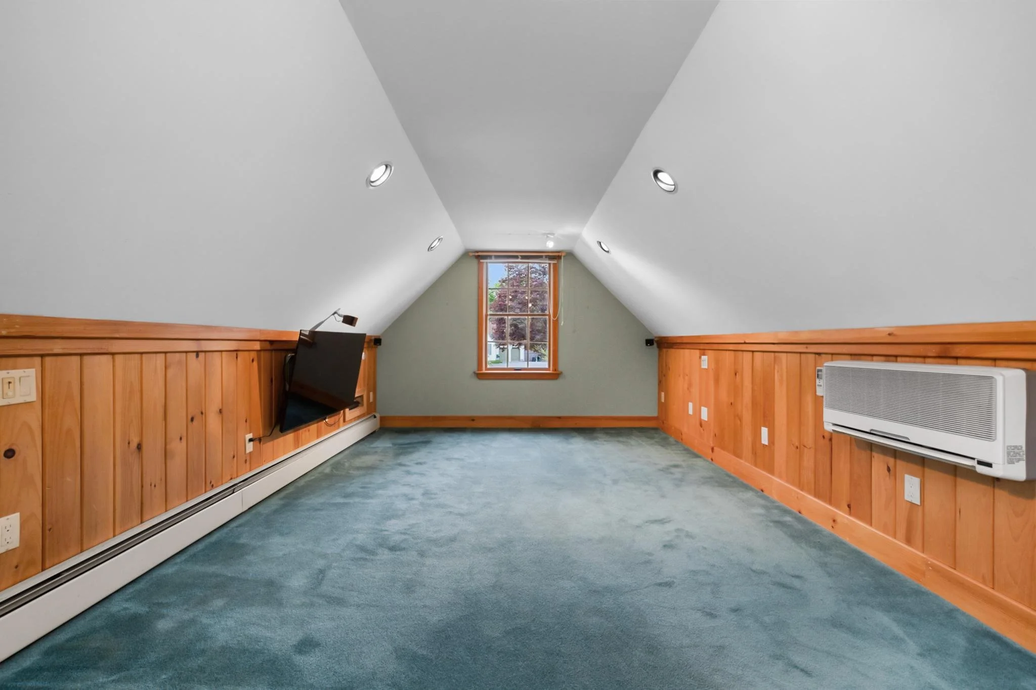 An empty room with sloped ceiling, white walls, and wood paneling. There are four ceiling lights, a window with a view of trees outside, a flat screen TV mounted on the wall, and a wall heater.