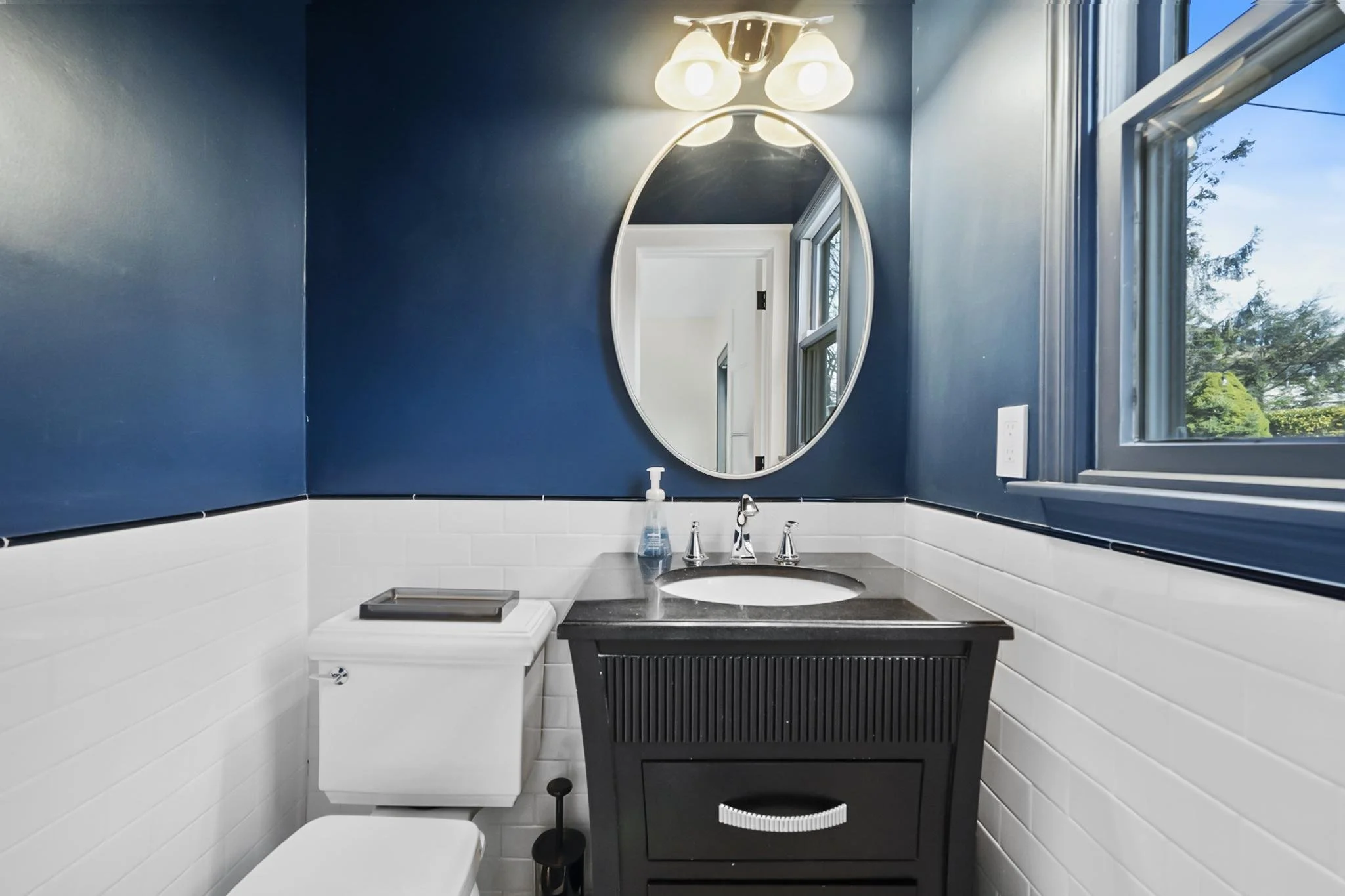 Bathroom with dark blue walls, oval mirror, window showing trees outside, and small black vanity with sink.