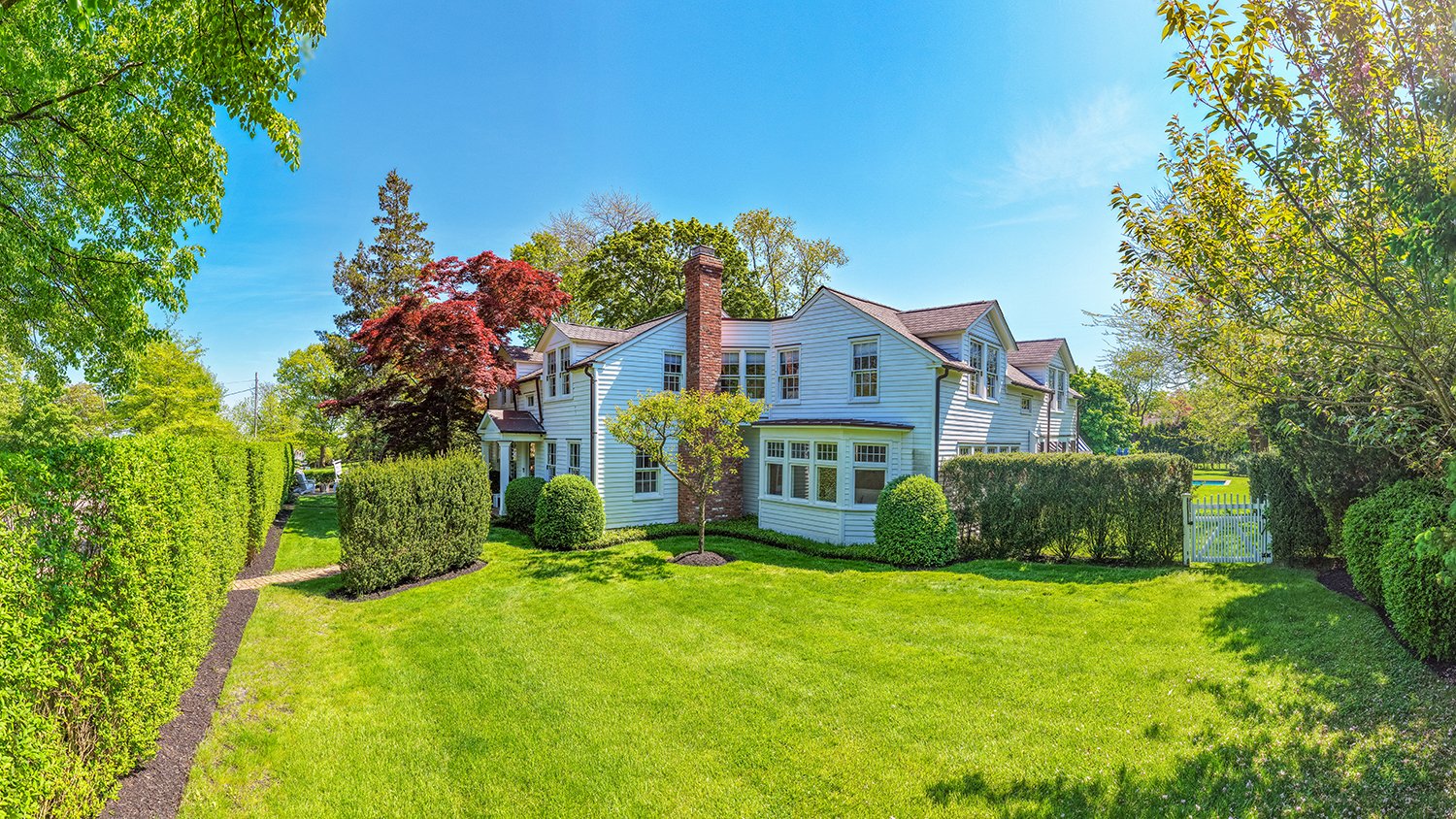 A large white house with multiple windows and a brick chimney, surrounded by green trees and a neatly maintained lawn under a bright blue sky.