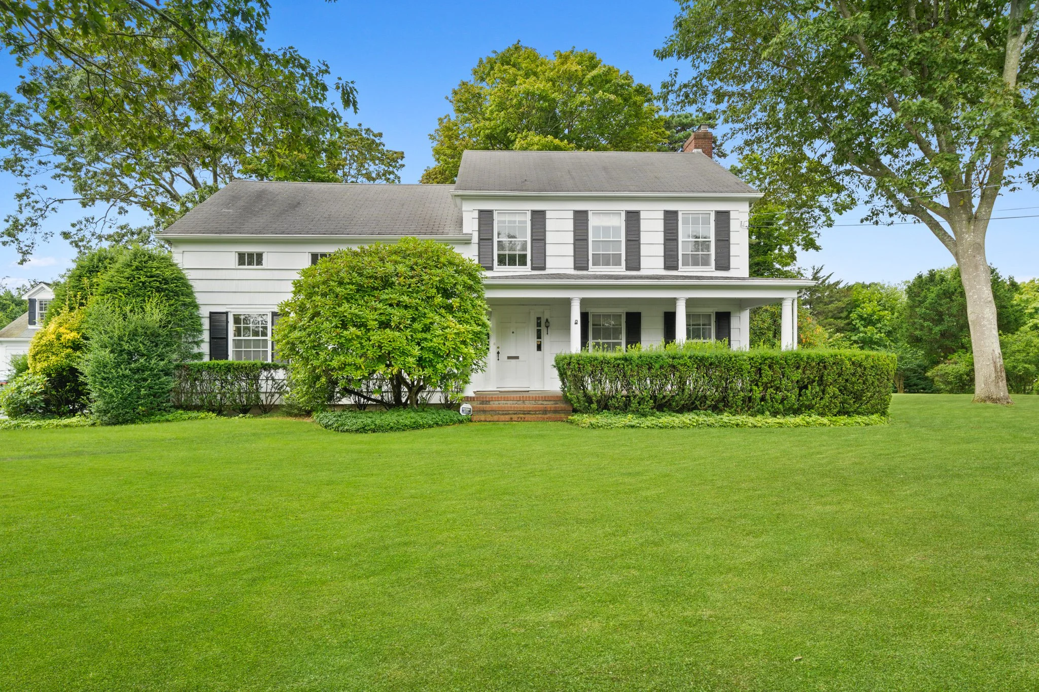 White two-story house with black shutters, a front porch supported by columns, and a well-maintained lawn with bushes and trees surrounding it.