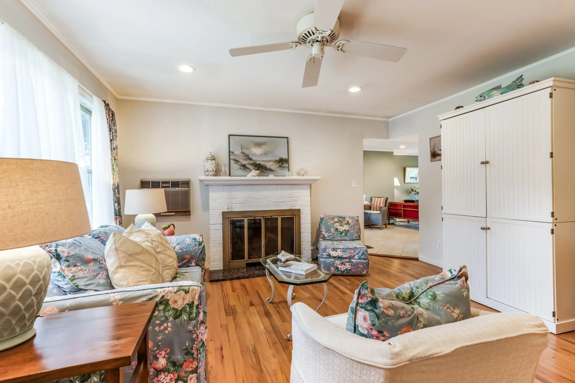 Living room with floral furniture, a white brick fireplace, and a ceiling fan. It has wood floors and natural light coming through large windows with white curtains.
