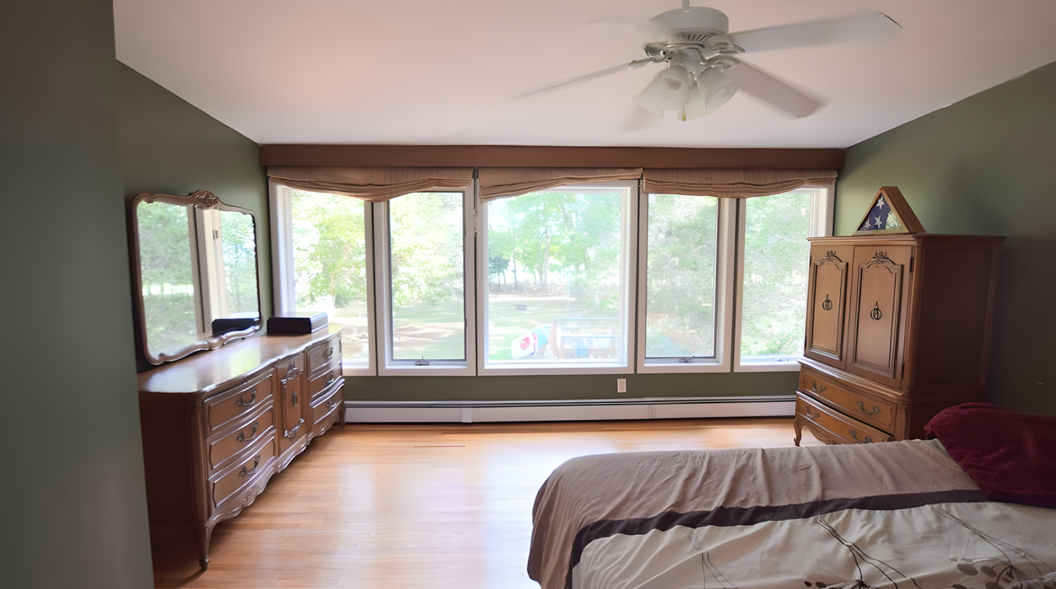 A bedroom with green walls, light wood flooring, large windows with brown valances, a ceiling fan, a wooden dresser with a mirror, a wooden armoire, and part of a bed with a beige and brown comforter and red pillow.