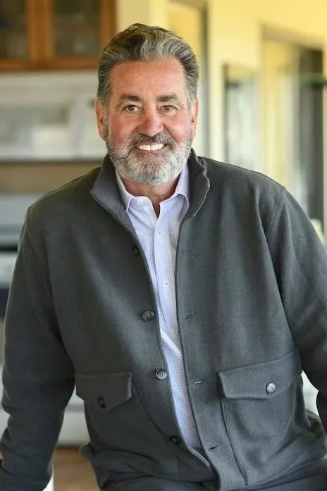 A smiling middle-aged man with gray hair and a beard, wearing a gray jacket and light blue collared shirt, standing inside a room with a kitchen in the background.