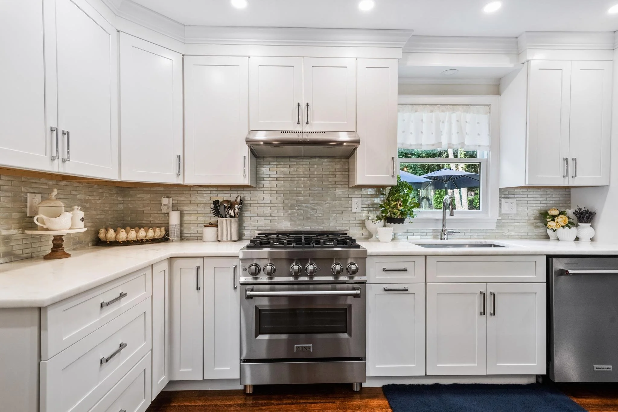 White kitchen with upper and lower cabinets, stainless steel stove and range hood, granite countertops, a window with curtains, and decorative plants and ceramics.
