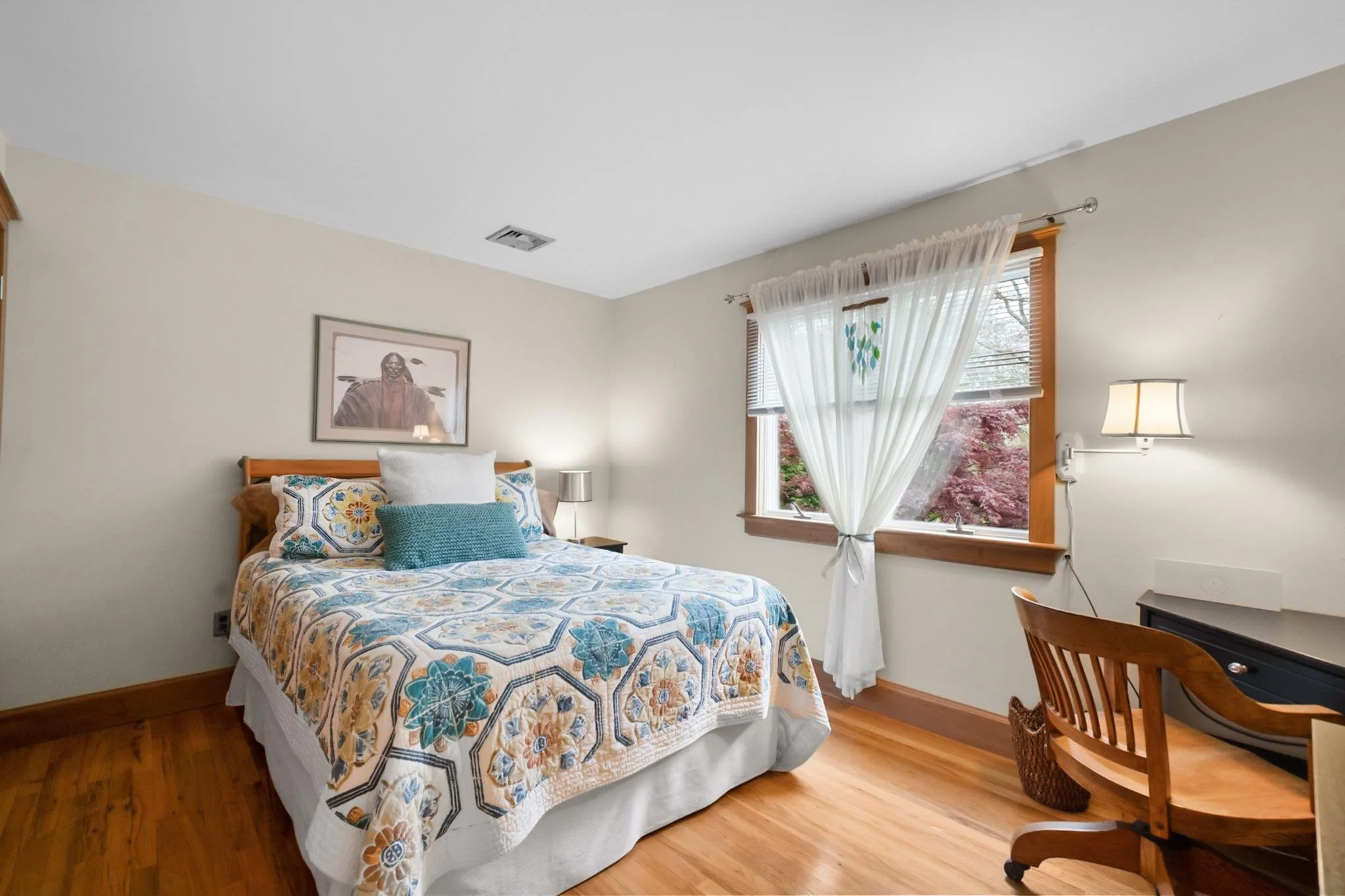 A cozy bedroom with a wooden bed frame, floral quilt, decorative pillows, a framed picture on the wall, window with white curtains, and wooden flooring.
