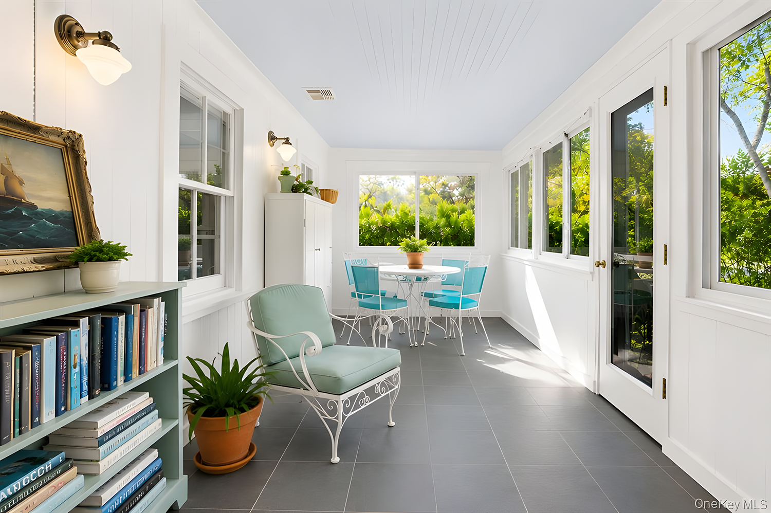 Bright, enclosed porch with white walls, large windows, gray tiled floor, a white round dining table with blue chairs, a white cabinet, a bookshelf with books, and potted plants, overlooking green foliage outside.