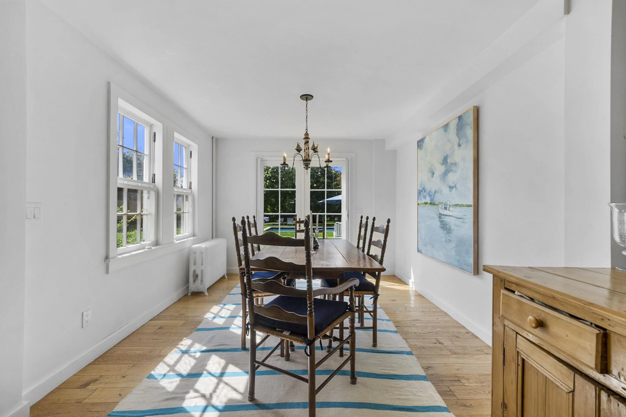 Dining room with wooden table, eight chairs, white walls, large windows, a colorful painting of a boat on water, chandelier, wooden sideboard, and hardwood floors.