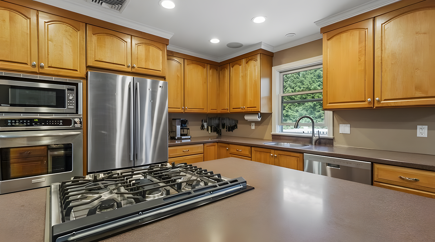 A modern kitchen with wooden cabinets, stainless steel appliances, and a window above the sink showing green trees outside.