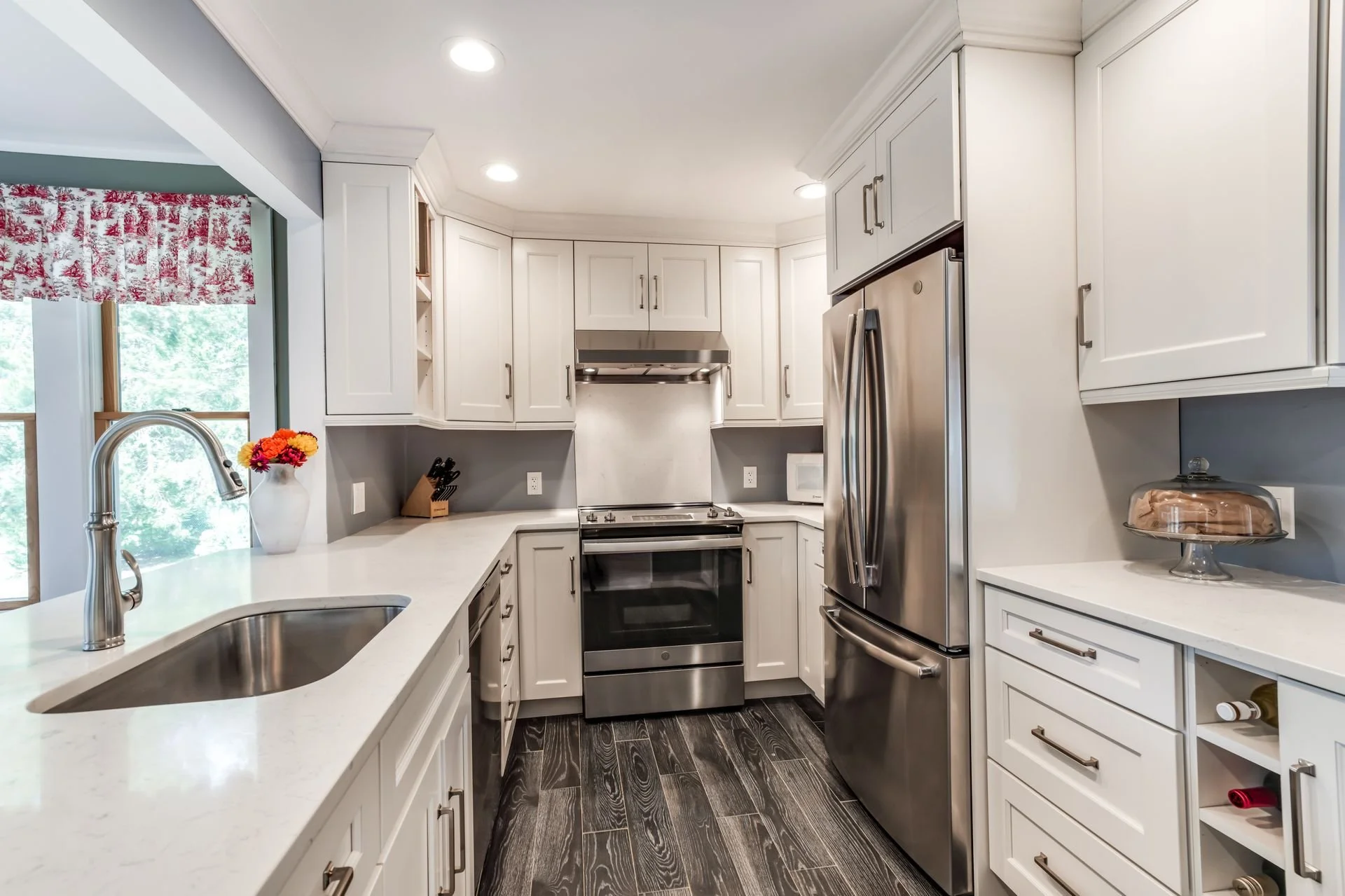 A modern kitchen with white cabinetry, stainless steel appliances, a black stove, and a window with a red and white patterned valance. There is a white countertop with a vase of flowers, a microwave, and a cake on a cake stand.