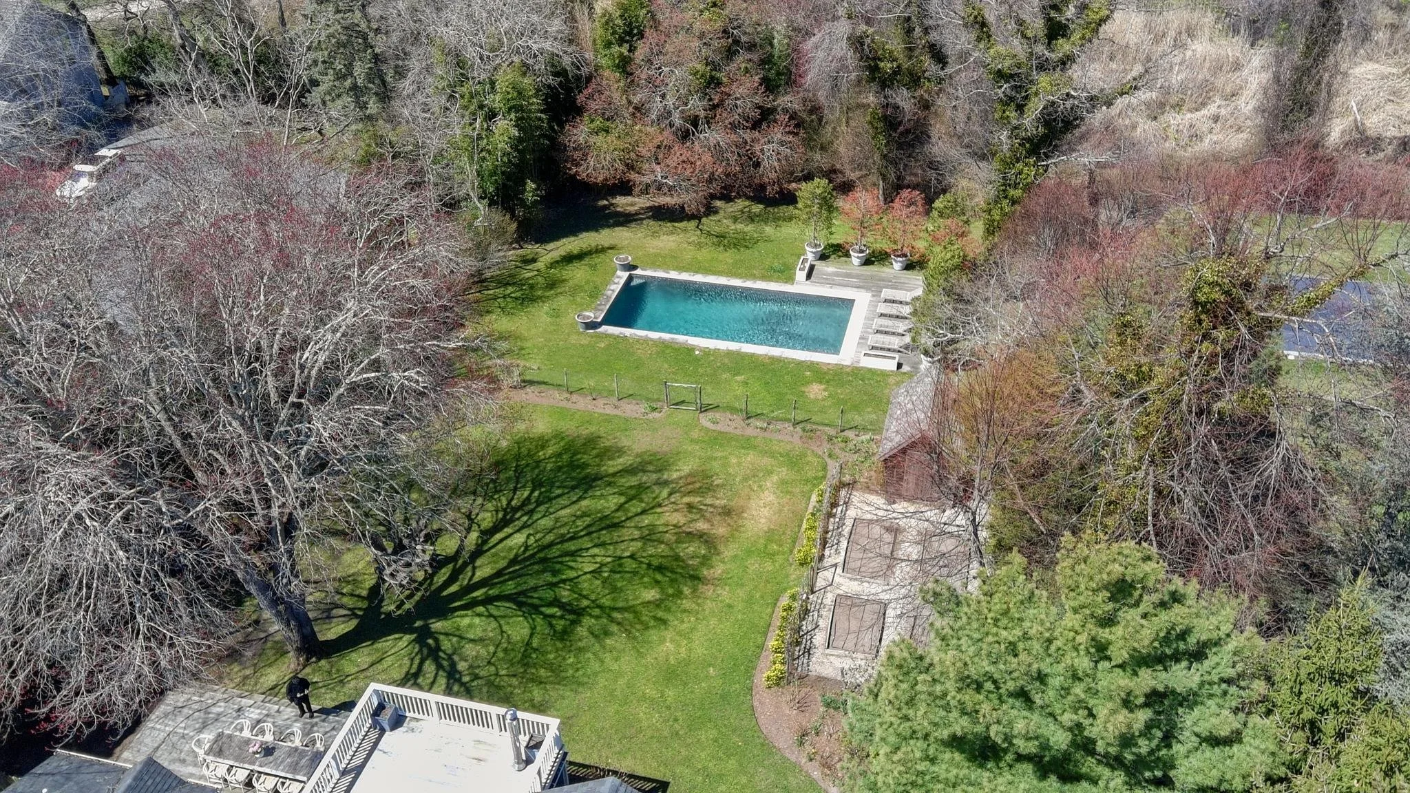 An aerial view of a backyard with a swimming pool, surrounded by trees, grass, and a patio area with lounge chairs.
