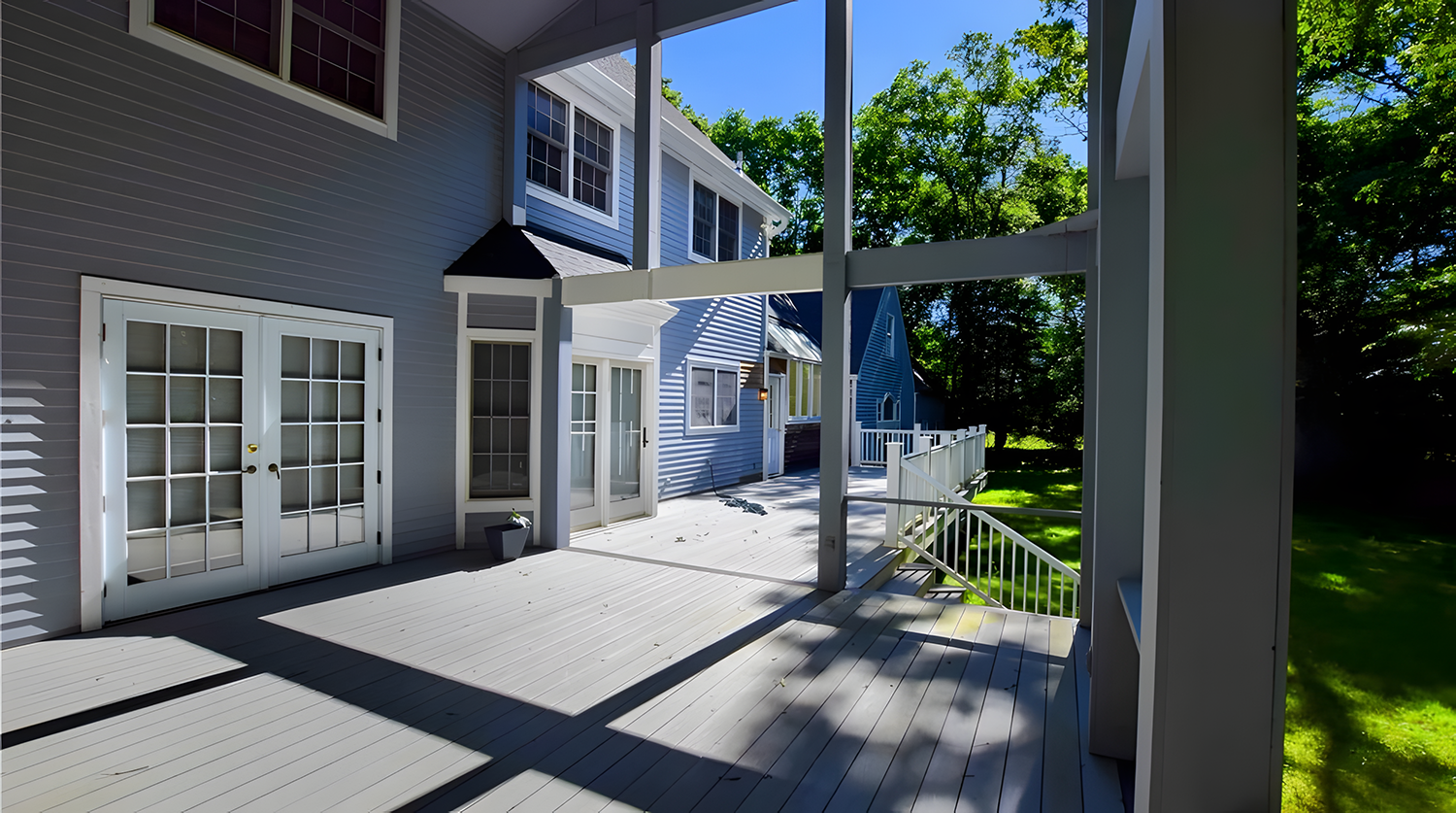 View of a backyard deck attached to a house, with French doors and windows, surrounded by green trees and grass, under a clear blue sky on a sunny day.
