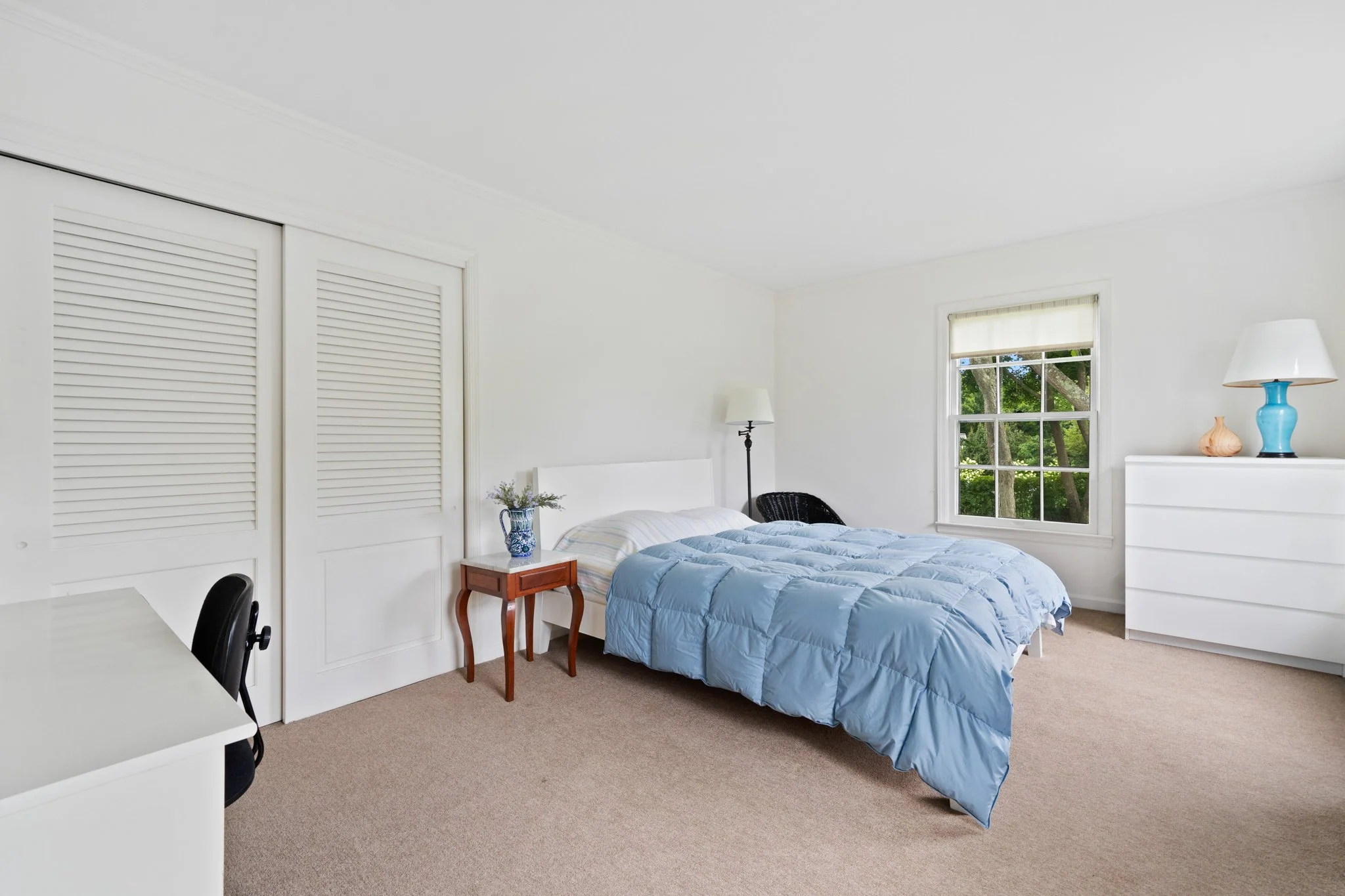 A bedroom with a bed covered in a light blue quilt, a small wooden side table with a blue vase and flowers, a white dresser with decorations, a window showing green trees outside, a black chair, and a white closet with louvered doors.