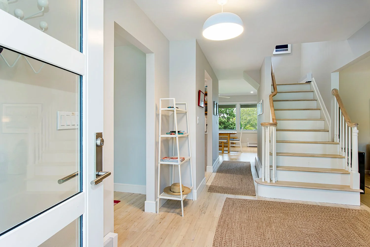 Entryway of a home with wooden stairs and a view into the dining area with large windows, light-colored walls, and hardwood floors.