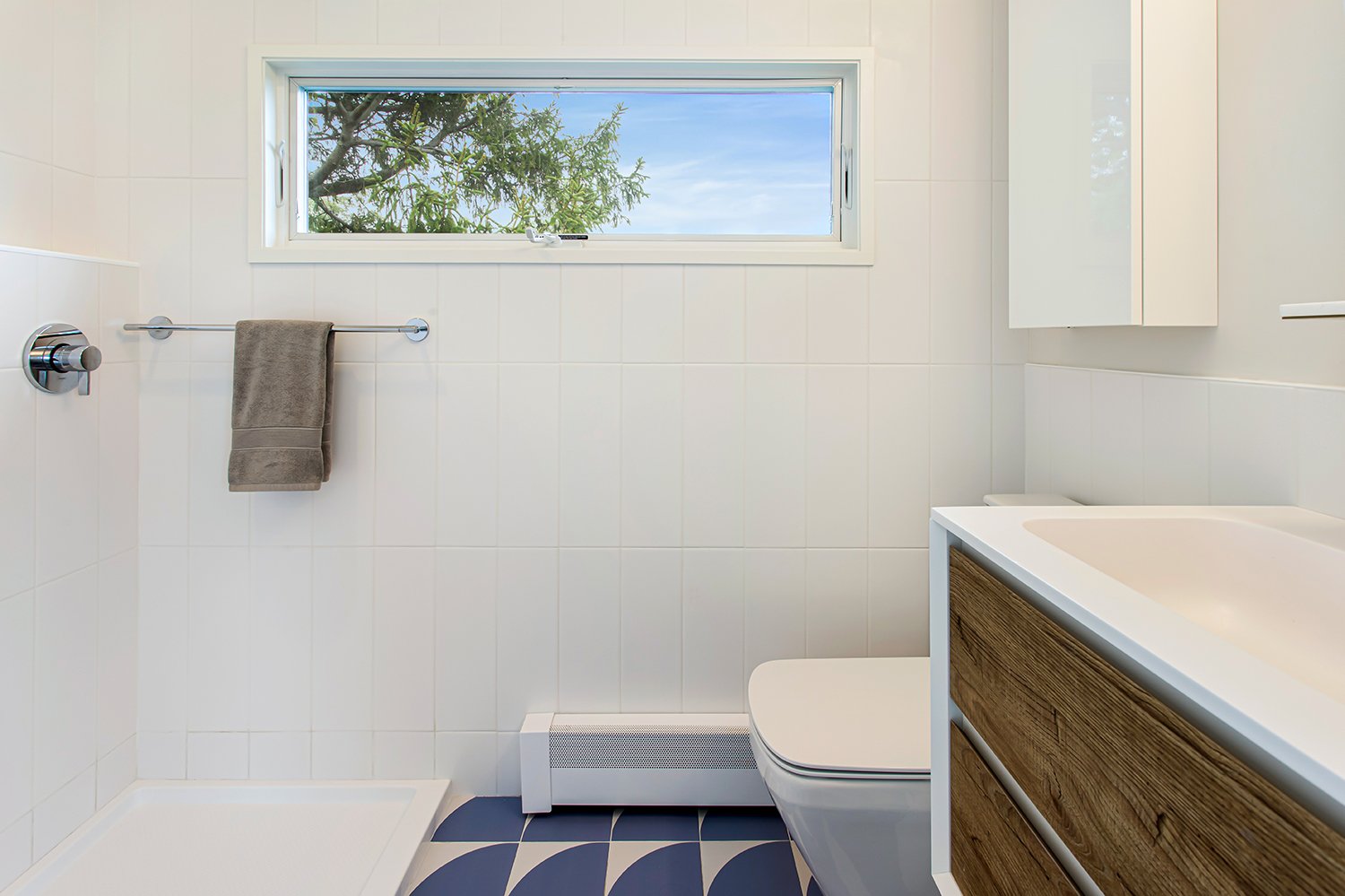 Modern white bathroom with a small window showing a blue sky and tree branches, featuring a walk-in shower, a towel on a chrome towel bar, a white toilet, and a wooden vanity with a sink.