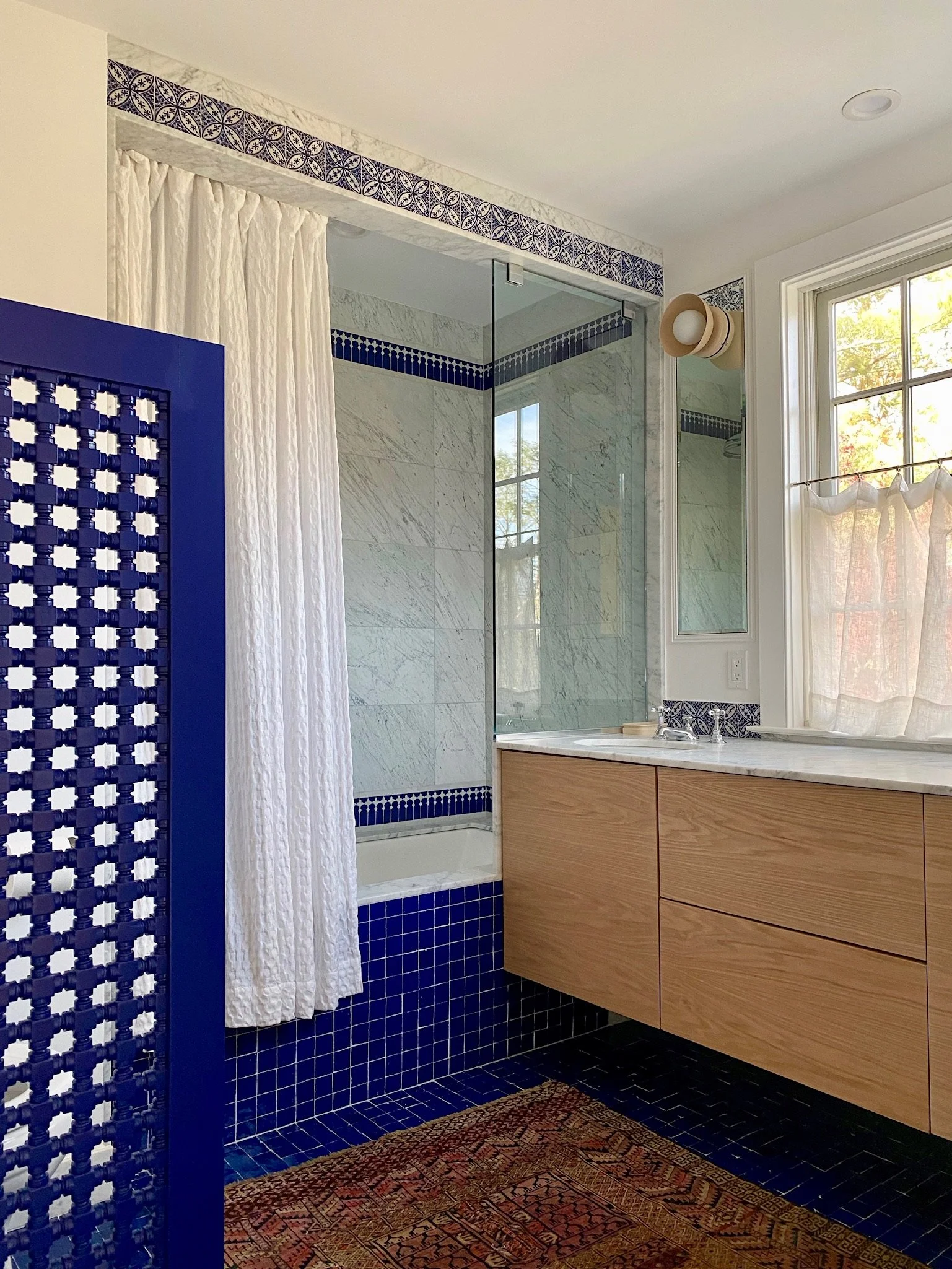 A bathroom featuring a walk-in shower with a glass door, white marble wall tiles with a decorative blue tile border, a wooden vanity with drawers, a large window allowing natural light, a blue partition with a lattice pattern, and a colorful patterne