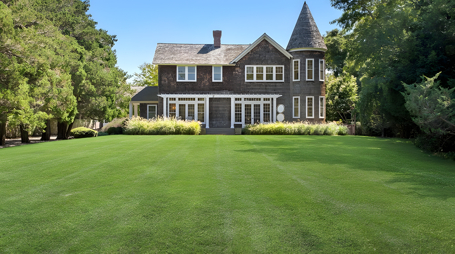 A large house with a turret and multiple windows, surrounded by a green lawn and trees under a clear blue sky.
