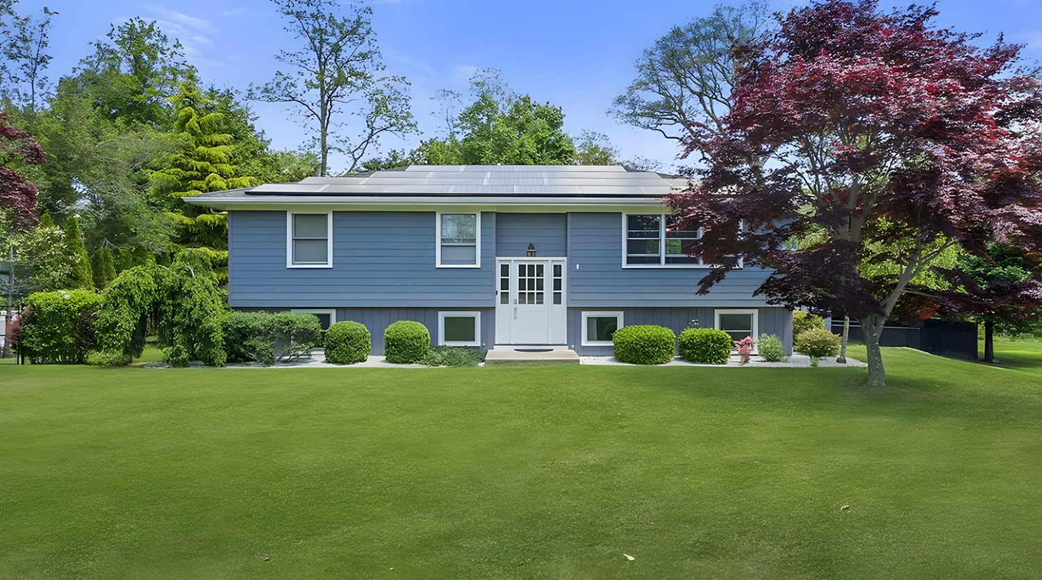 Blue house with solar panels on the roof, surrounded by green lawn and trees, including a large red-leafed tree on the right side