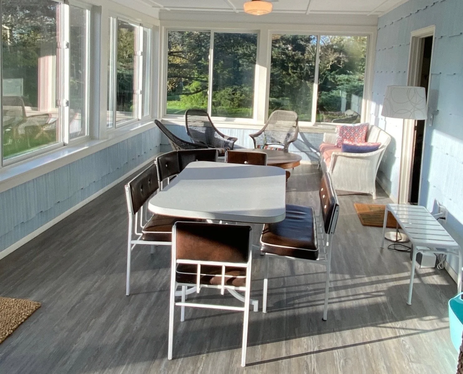 Sunlit enclosed porch or sunroom with large windows, containing a white rectangular table surrounded by black and brown chairs, along with a white wicker sofa with colorful cushions, a white floor lamp, a small white side table, a wicker basket, and 