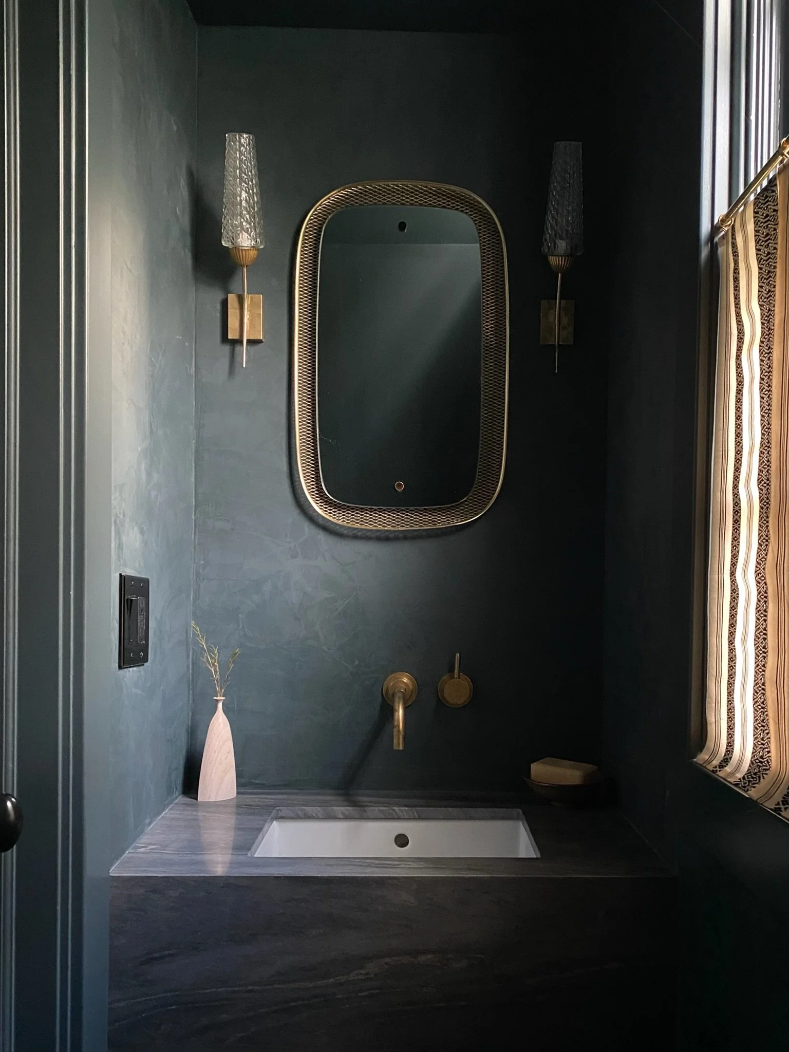 A modern bathroom with dark green walls, a rectangular white sink, a large oval mirror with a gold frame, two gold wall sconces with black glass shades, a beige vase with a plant, and a shower area with gold fixtures.