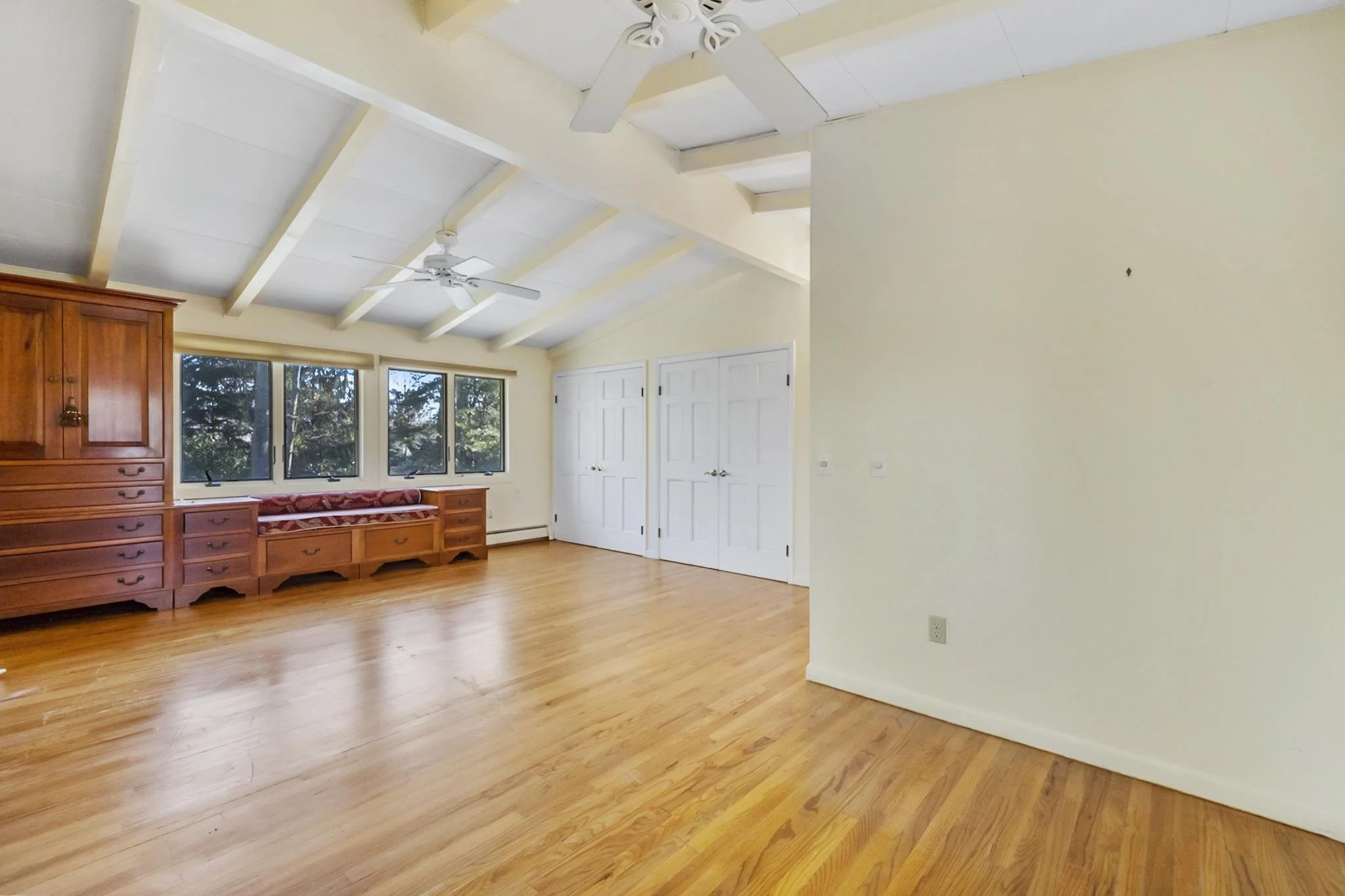 A bright living room with large windows, wooden floors, a wooden cabinet, white double doors, and ceiling fans.