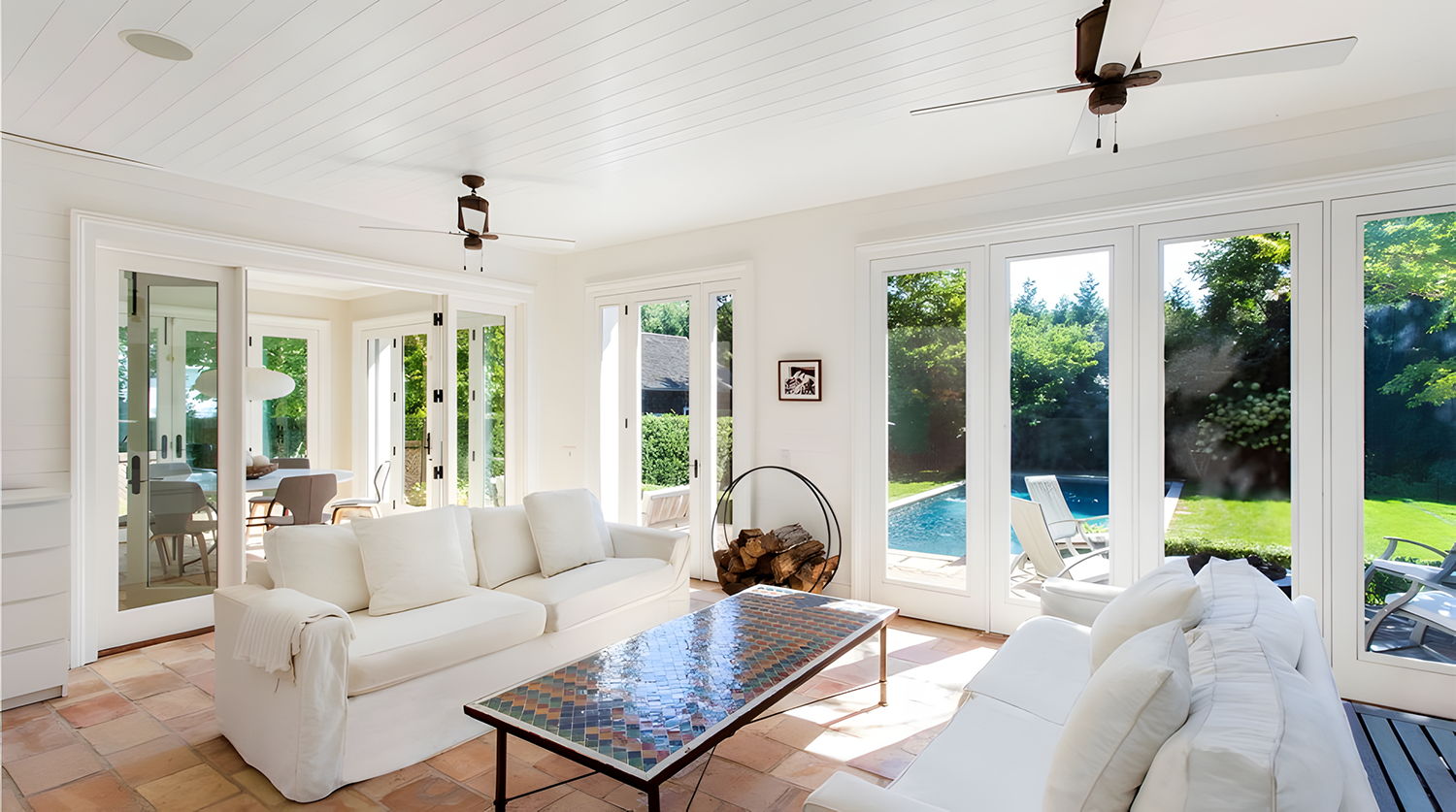 Bright living room with white sofas and a mosaic coffee table, glass doors leading to a backyard with a swimming pool and outdoor chairs, and a view of greenery outside.