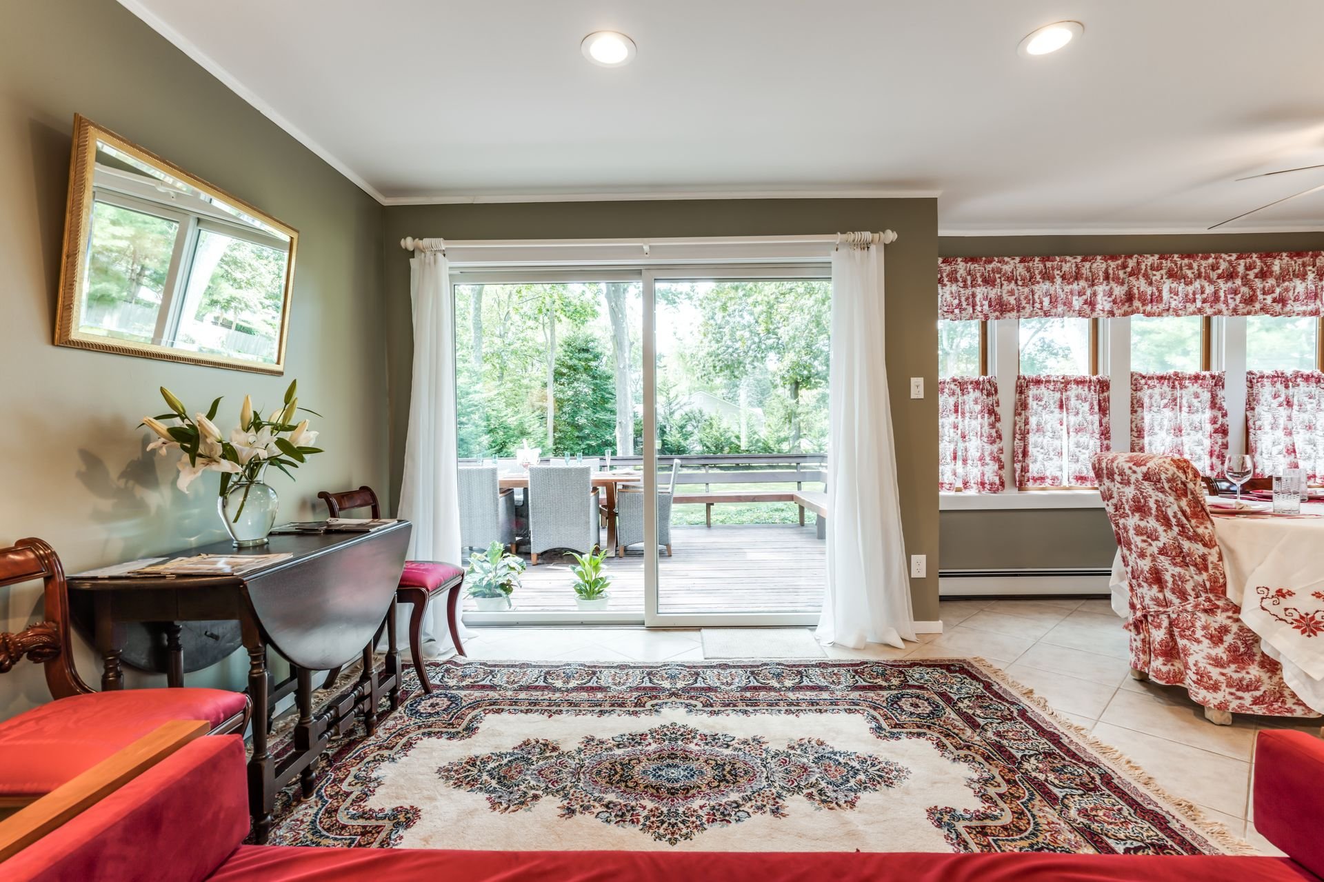 Living room with sliding glass door leading to patio, decorated with curtains and floral-patterned window valance, sofa, dining table, and chairs, illuminated by natural light.